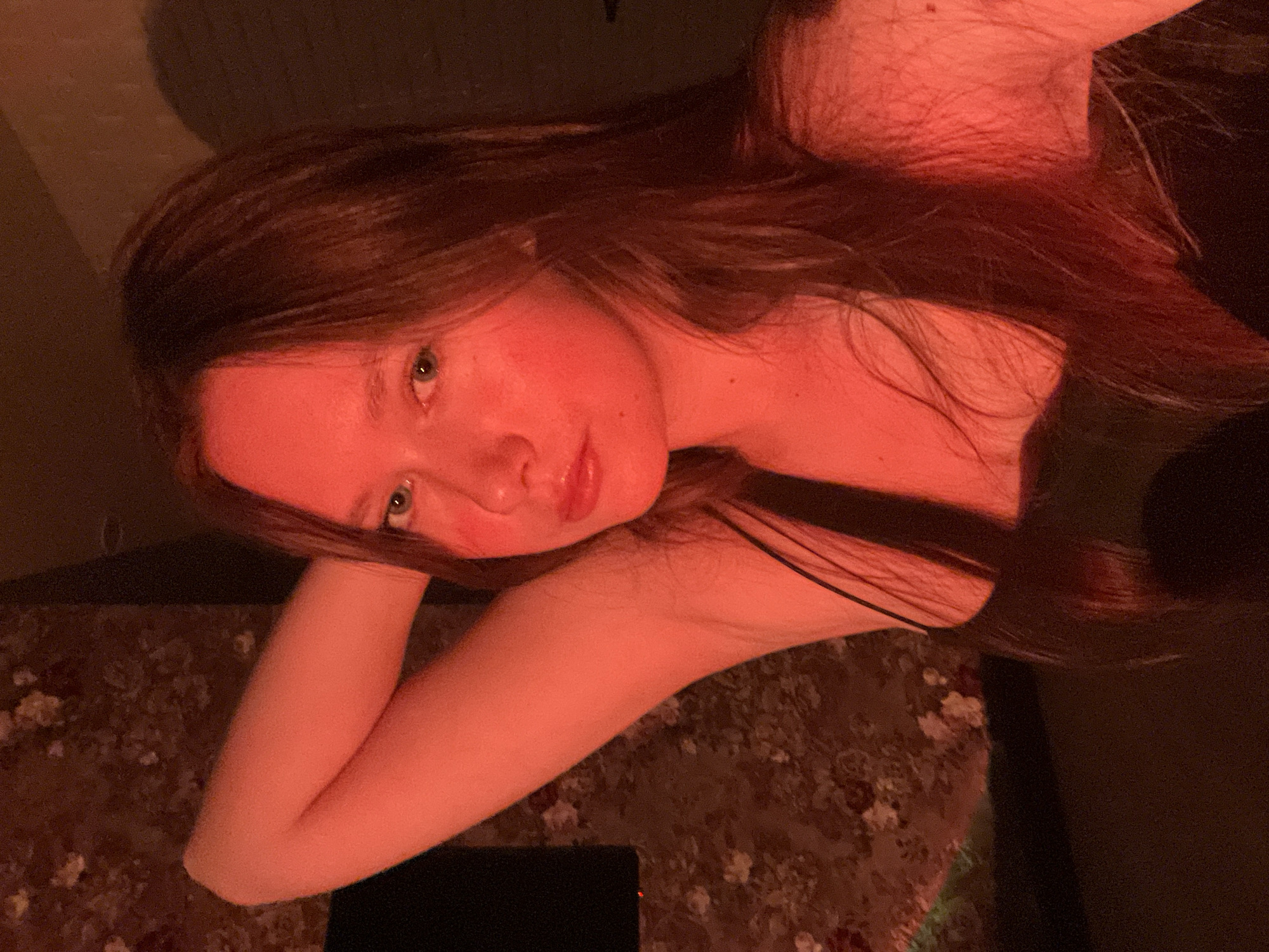 The image features a young woman with long, dark brown hair, captured in a close-up portrait. Her fair skin is illuminated by a strong, warm, red-orange light, casting a reddish hue over her complexion and hair. She is wearing a dark, possibly black, spaghetti strap top or dress. The woman gazes directly at the camera with a soft, slightly pensive expression, her head tilted. Her left arm is bent, and her hand gently supports her head near her chin, while her right arm rests alongside her body.
The setting appears to be indoors, likely in the evening or night, given the low, artificial red lighting that dominates the scene and creates an intimate atmosphere. The background is dimly lit; to her left, a dark, indistinct surface is visible, possibly a wall. To her right and below her arm, there is a patterned surface, resembling a carpet or upholstered furniture, with faded floral or abstract designs in muted earthy tones. A faint rectangular glow, possibly from a mobile device screen, is reflected on this surface near her right elbow. No visible text is present. The provided location context for the image is Chaikovsky, Russia.