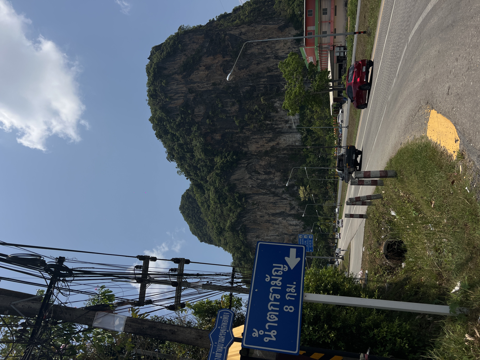 This is an outdoor photograph of a road scene in Tambon Takdat, Thailand. In the background is a large, rocky mountain covered with green vegetation. A two-lane road runs alongside the mountain, with a few vehicles including a red car and a dark-colored SUV on the road. Utility poles with tangled wires are visible on the left side of the frame.
A blue road sign in the foreground has white text in Thai script. Some of the text on the sign says "น้ำตกรามัญ 8 กม.", which translates to "Raman Waterfall 8 km". The sign also has a left-pointing arrow. Additional sign details are shown in the bottom left corner.
The weather appears to be sunny with a blue sky and scattered white clouds. The roadside is bordered by a grassy area with litter and a few small concrete barriers.
