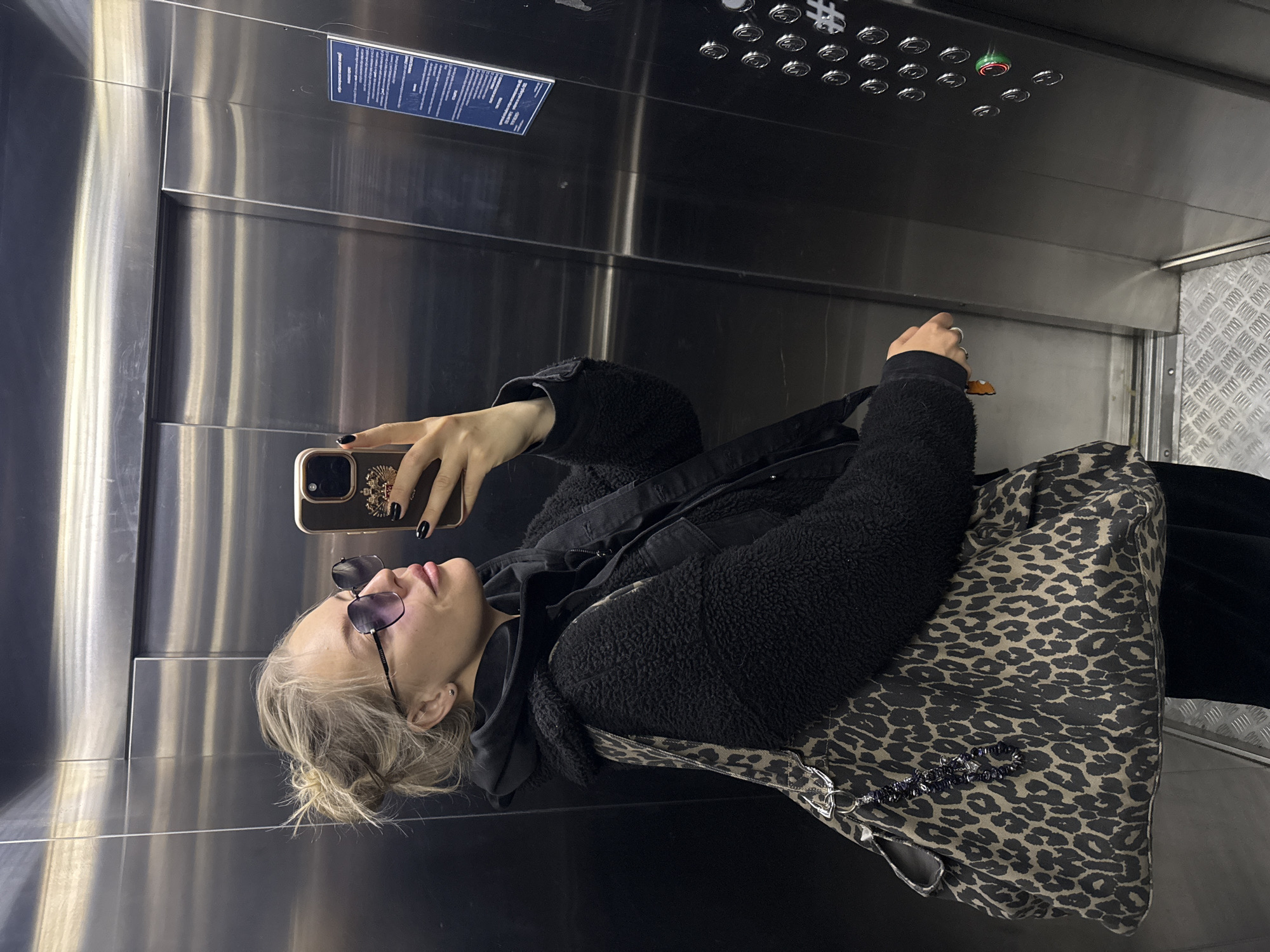 A woman is taking a selfie in an elevator. The elevator has a brushed steel interior, with a panel of buttons and a sign at the top. The woman is wearing sunglasses, a black jacket, a hoodie, and a leopard print bag. She is holding a cell phone up to take the picture. Based on the available information, it's not possible to confirm that this photograph was taken in Krasnoyarsk, Russia.