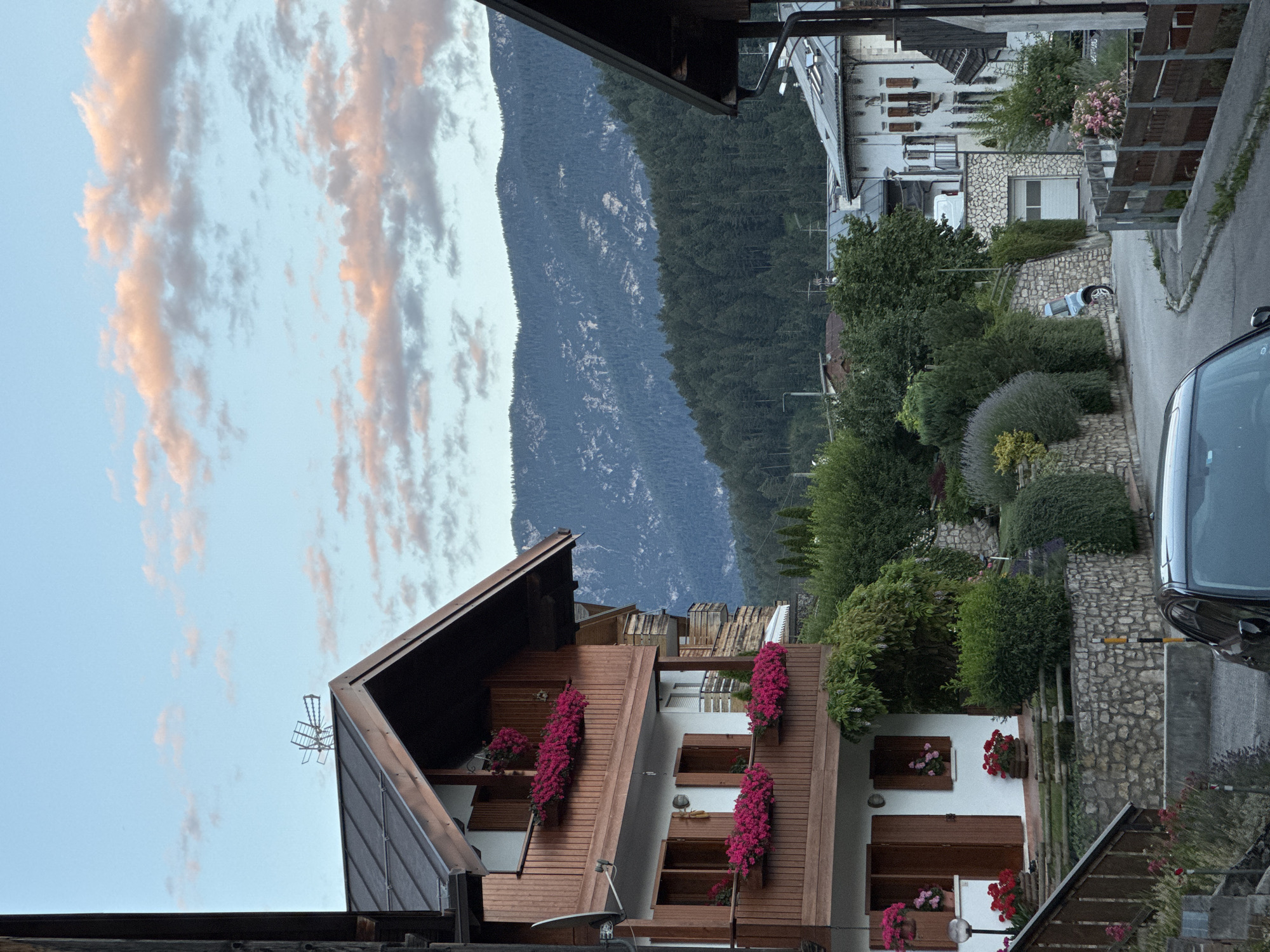The image depicts a street scene in Valle di Cadore, Italy, at dusk. A multi-story building with wooden accents and balconies adorned with vibrant pink flowers dominates the left side of the frame. The building has white walls, dark wooden shutters, and a dark roof. In the background, lush green trees cover the slopes of a mountain range under a sky with scattered pink-tinged clouds. A paved road curves through the scene, with a dark-colored car parked on the right and a light blue scooter leaning against a stone wall on the left. Other residential buildings and greenery are visible further down the street. The overall impression is a serene, picturesque mountain town at the end of the day.