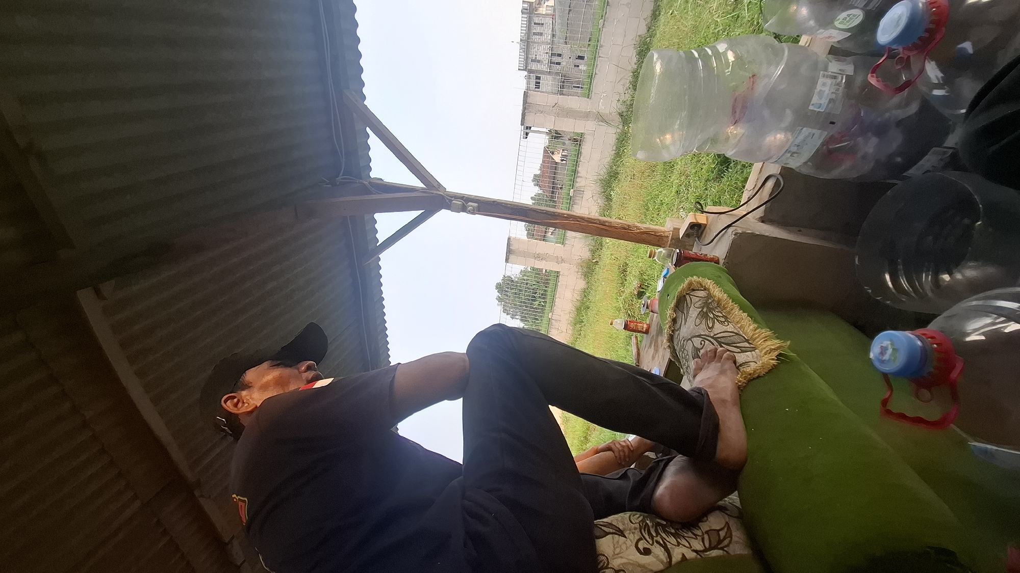 The image depicts a person wearing a black t-shirt and black pants, seated on a green sofa under a corrugated metal roof. The person is wearing a black baseball cap and is looking away from the camera. In the foreground, several large plastic bottles are visible. Beyond the structure, a fence separates the area from a grassy field with some buildings in the distance. The scene appears to be outdoors in Rahong, Indonesia.