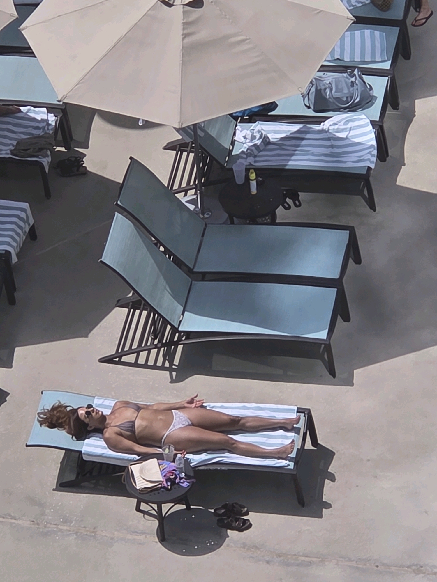 A woman is sunbathing on a lounge chair on a patio in Long Beach, California, USA. She is wearing a bikini and sunglasses. The lounge chair is on a striped towel. A small round table is next to the chair, with a drink and a book on it. In the background, there are other lounge chairs and umbrellas. The sun is shining and casting shadows on the ground.