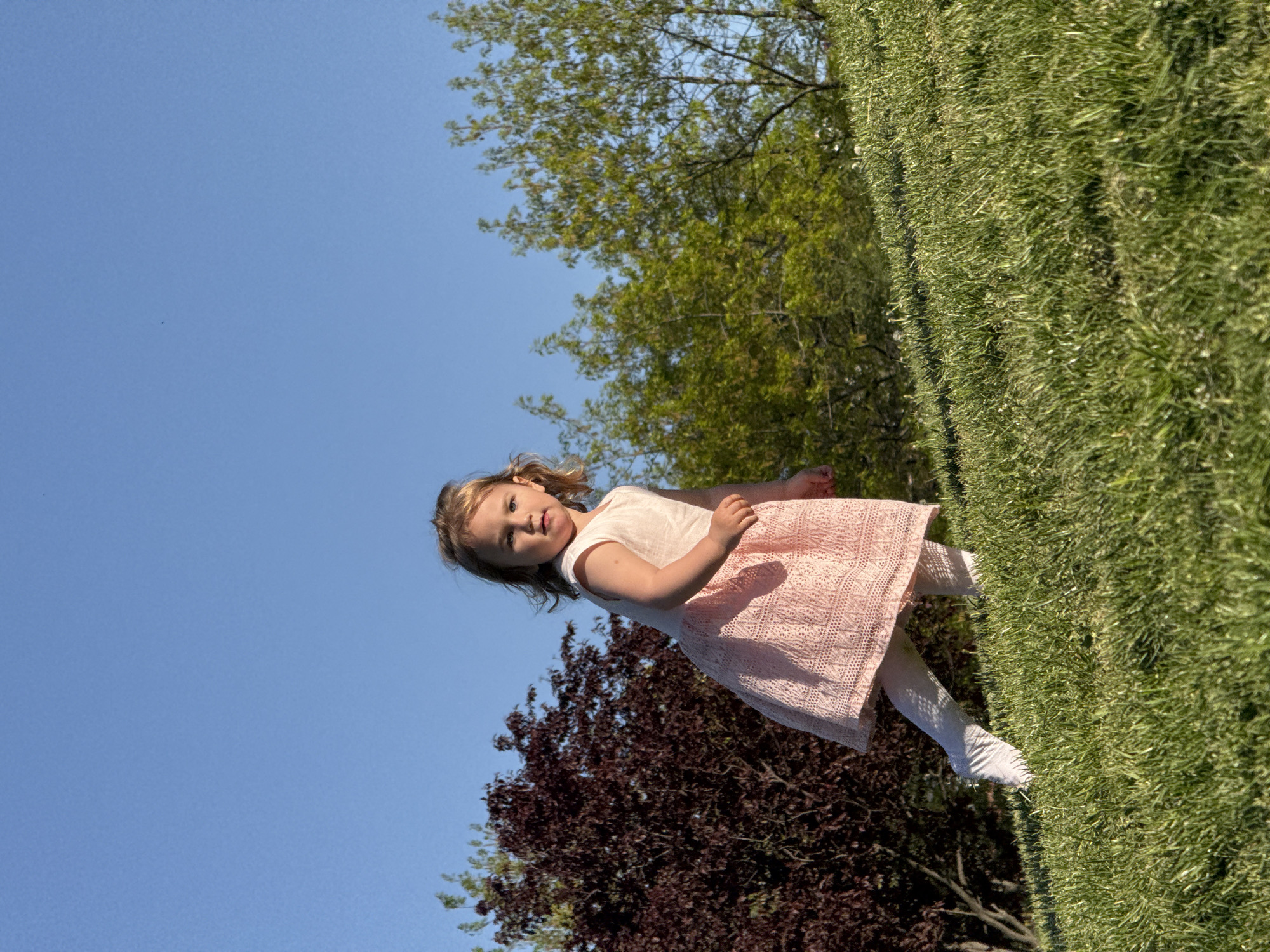 A young girl in a peach-colored dress and white tights stands in a grassy area outdoors. Behind her are trees with green and maroon foliage, and the sky is a clear blue. This scene is likely not in Sofia, Bulgaria as there is no information to confirm it.