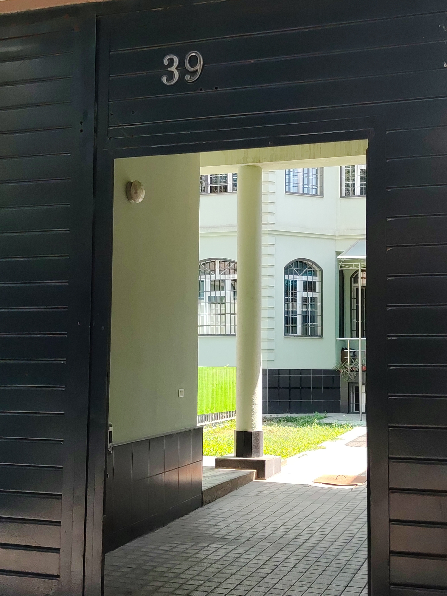 An exterior view captures the entrance to a building, identified by the number "39" prominently displayed above. The entrance is framed by dark, horizontal slatted doors, partially open to reveal a courtyard. The courtyard features a paved walkway with a geometric pattern, leading towards a light-colored building with multiple windows. The building's facade is a pale green, and its windows are divided into geometric panes. A substantial cylindrical column, topped with a decorative capital, stands to the left of the entrance, dividing the view into sections. The environment appears to be a sunny day, with shadows cast across the paving stones. The image provides a clear, static view of the building's entrance and its immediate surroundings, without any human presence or activity. The setting suggests a residential or administrative building within an urban area.