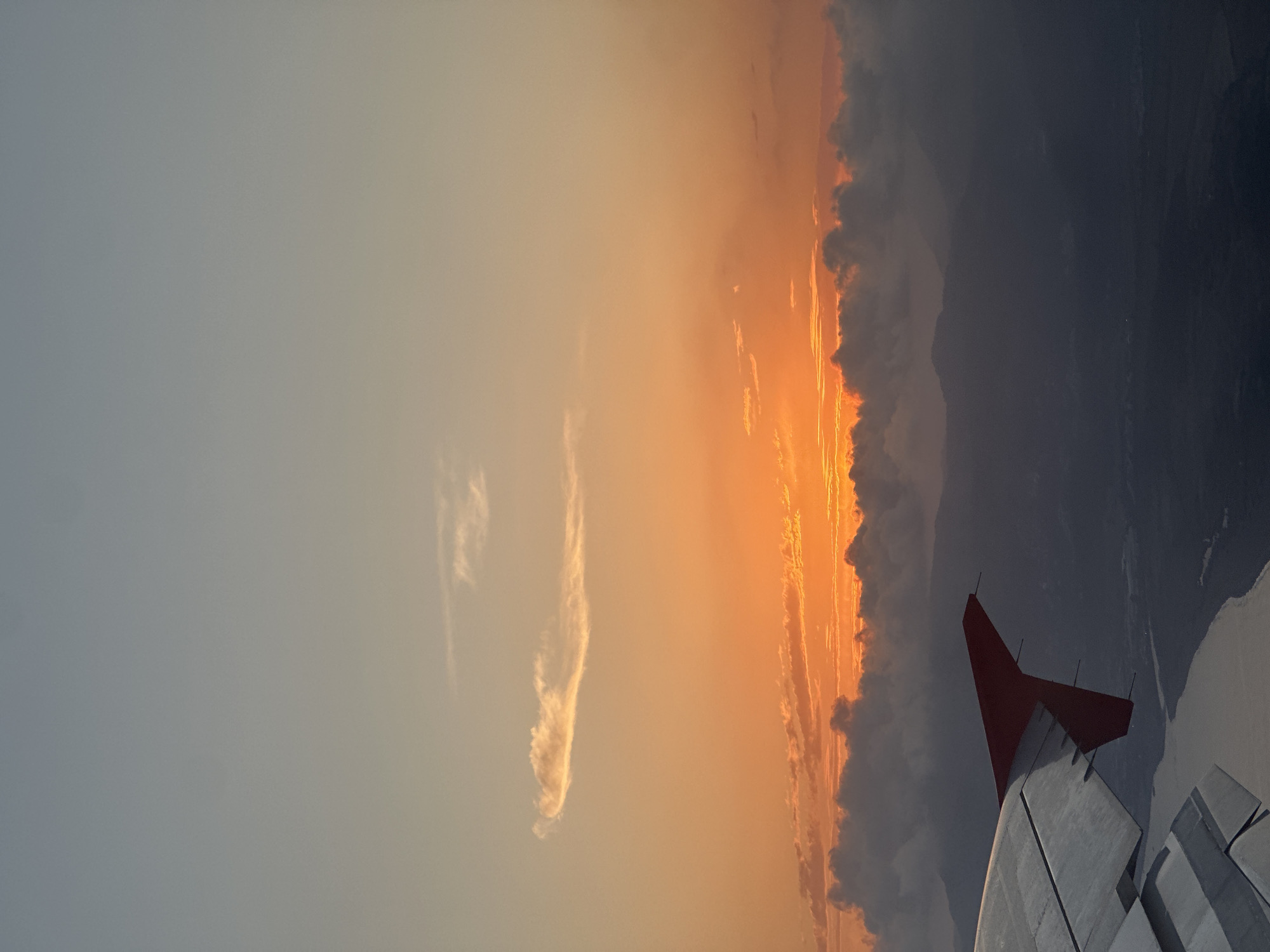 An aerial view from an aircraft captures a vivid sunset over a mountainous terrain near Dalaman, Turkey. The sky transitions from a pale blue-gray at the top, through warm orange mid-tones, to an intense, fiery orange glow at the horizon, where the sun is setting. A prominent, elongated cloud formation is illuminated by the sunset in the upper-mid section of the sky, with denser, dark clouds obscuring the lower portion of the sun's disc along the horizon. The left wing of the aircraft, rendered in silver-gray with visible panels and trailing edge devices, dominates the lower-left quadrant, featuring a distinct red wingtip. Below the wing, a light-toned body of water, likely a river, curves through a dark, shadowed landscape characterized by undulating hills and distant mountains, which appear silhouetted against the bright horizon. No people are visible.