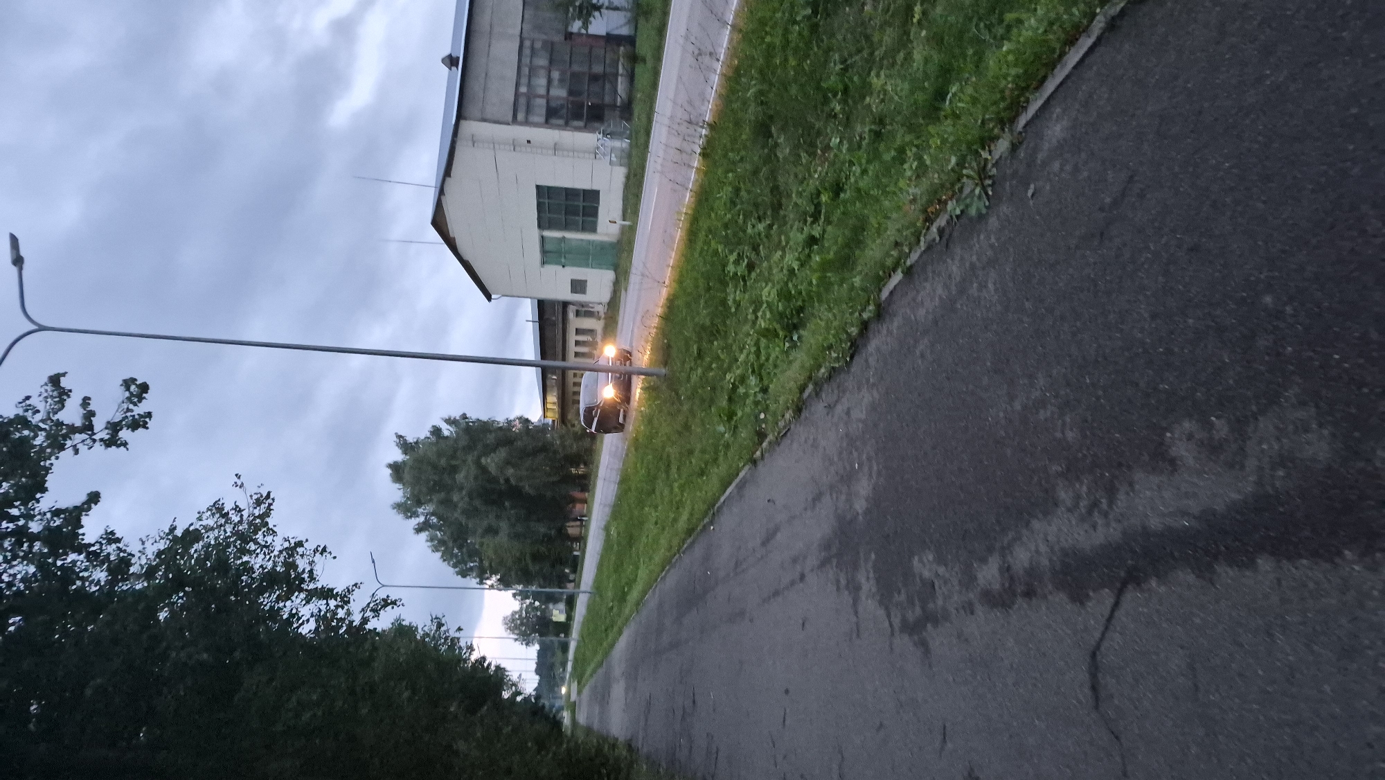 The image depicts a twilight scene along a paved walkway adjacent to a road in Krauja, Latvia. On the right side of the road, a dark-colored car with its headlights on is in motion, approaching the viewer. Beyond the road, a long, low-rise industrial building with a light-colored facade and a large green door is visible. Further in the background, more industrial structures with dark roofs are present. Trees line the left side of the walkway and the area bordering the road, their foliage dark against the overcast sky. Streetlights are visible, one prominently in the foreground and another further down the road. The sky is cloudy, suggesting either early morning or late evening. The ground of the walkway appears damp and somewhat cracked.