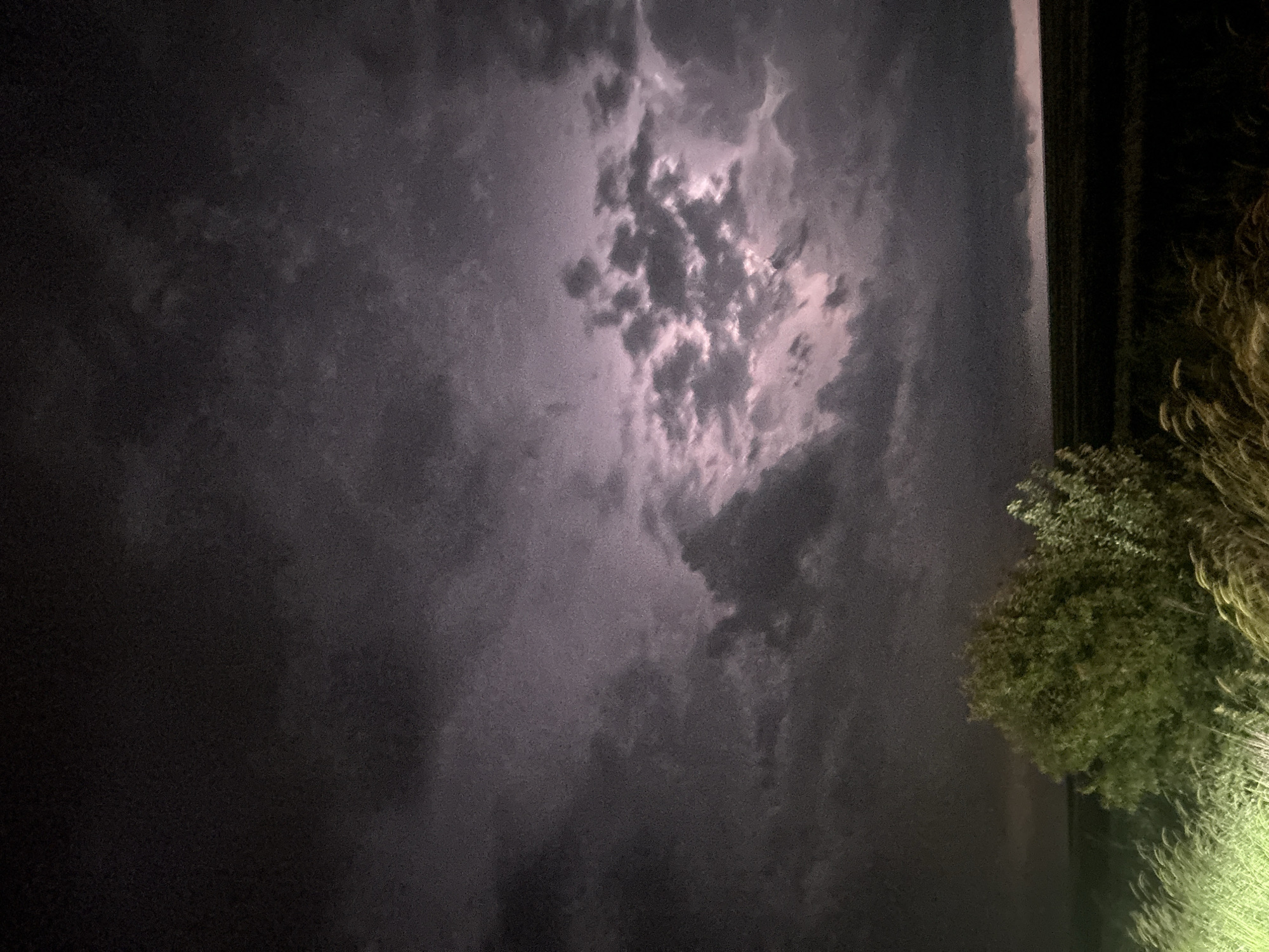 The image shows a dark, cloudy sky. To the right, there are illuminated trees and grass. A dark structure is also visible on the right side. The location in Burgas, Bulgaria is not discernible from the image.