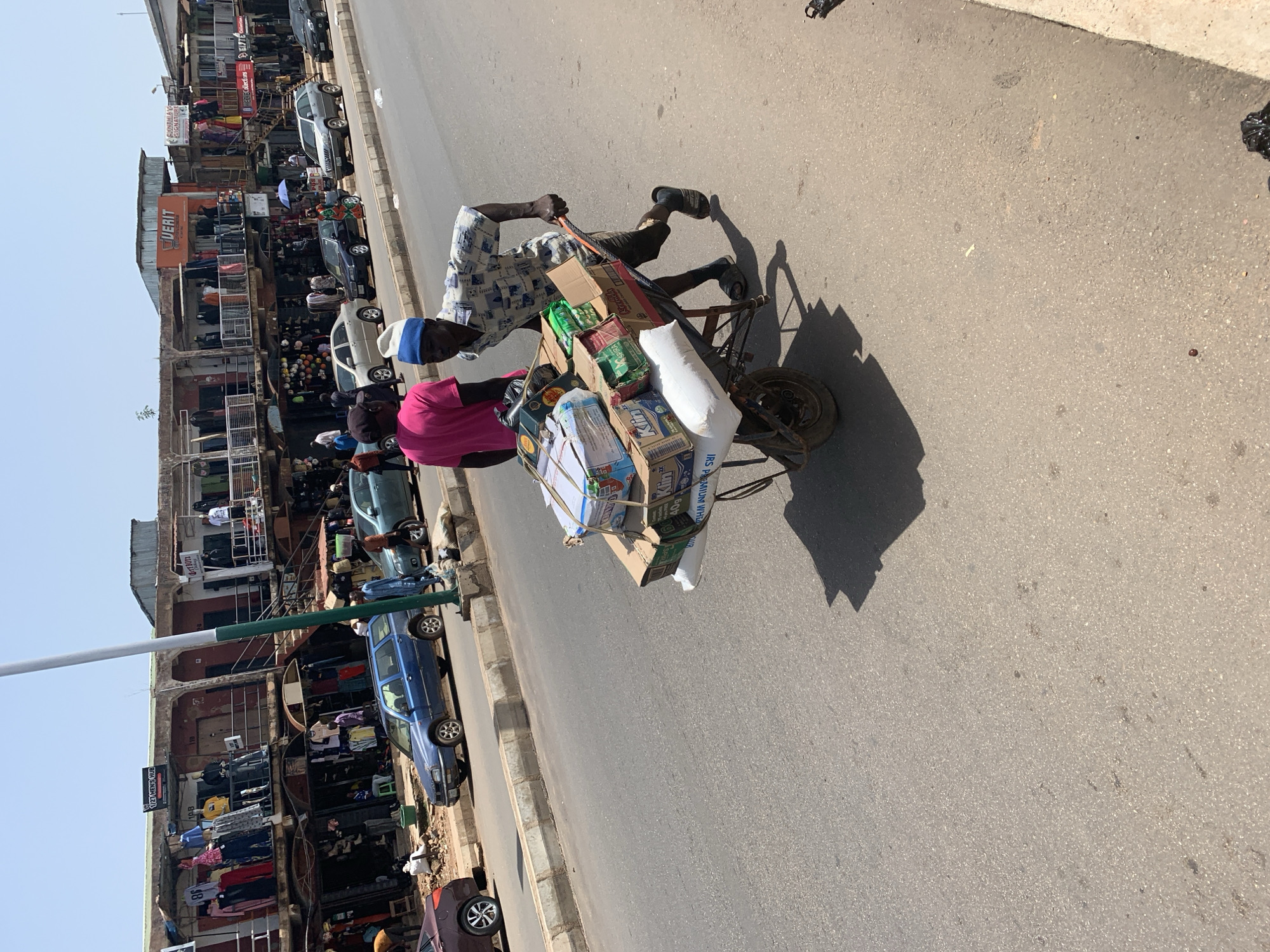 A man pushes a wheelbarrow loaded with boxes and bags of goods down a paved street in Jos, Nigeria. The man, wearing a patterned shirt and shorts, and a blue and white cap, is walking on the right side of the street. The wheelbarrow is piled high with various items, including what appear to be cartons of drinks, boxes of packaged goods, and large white sacks. In the background, a two-story building with shops and balconies lines the street. Several cars, including a blue SUV and a silver sedan, are parked along the curb. Another man, wearing a bright pink shirt, stands behind the man with the wheelbarrow. The scene appears to be a busy market area.