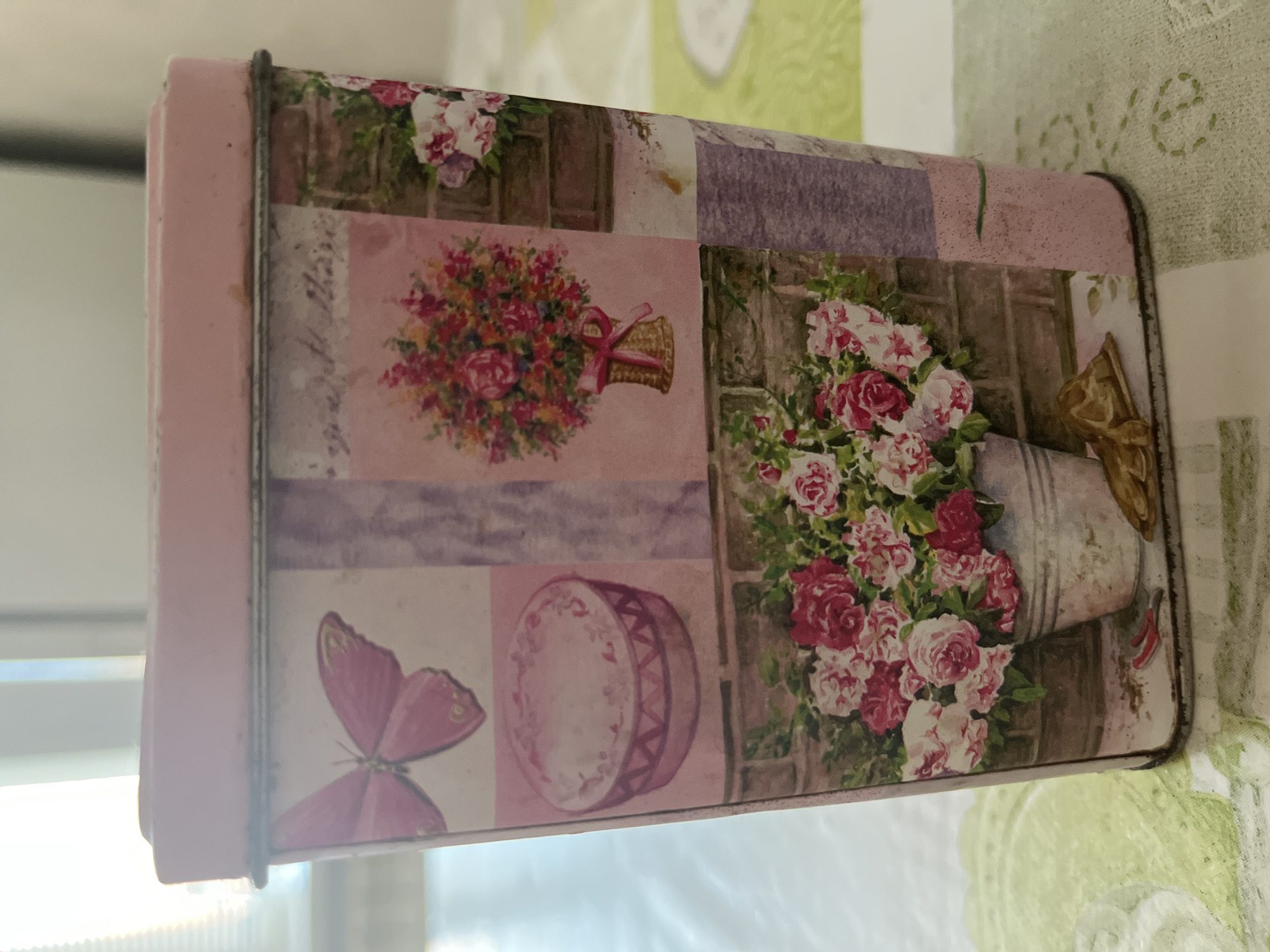 The image shows a close-up of a pink tin box with a decorative floral and butterfly design. The tin is rectangular and stands upright on a light green surface, possibly a tablecloth, with a subtle embroidered pattern. The design on the tin features various elements arranged in a collage-like fashion: a pink butterfly, a round pink object resembling a cake or hat box, a bouquet of colorful flowers in a woven basket tied with a pink ribbon, and a large arrangement of pink and white roses in a silver bucket. The background of the tin's design includes elements of brickwork and textured panels. The lighting suggests an indoor setting, with light streaming in from the left, possibly from a window. The tin appears to be a decorative container. The location is unknown.