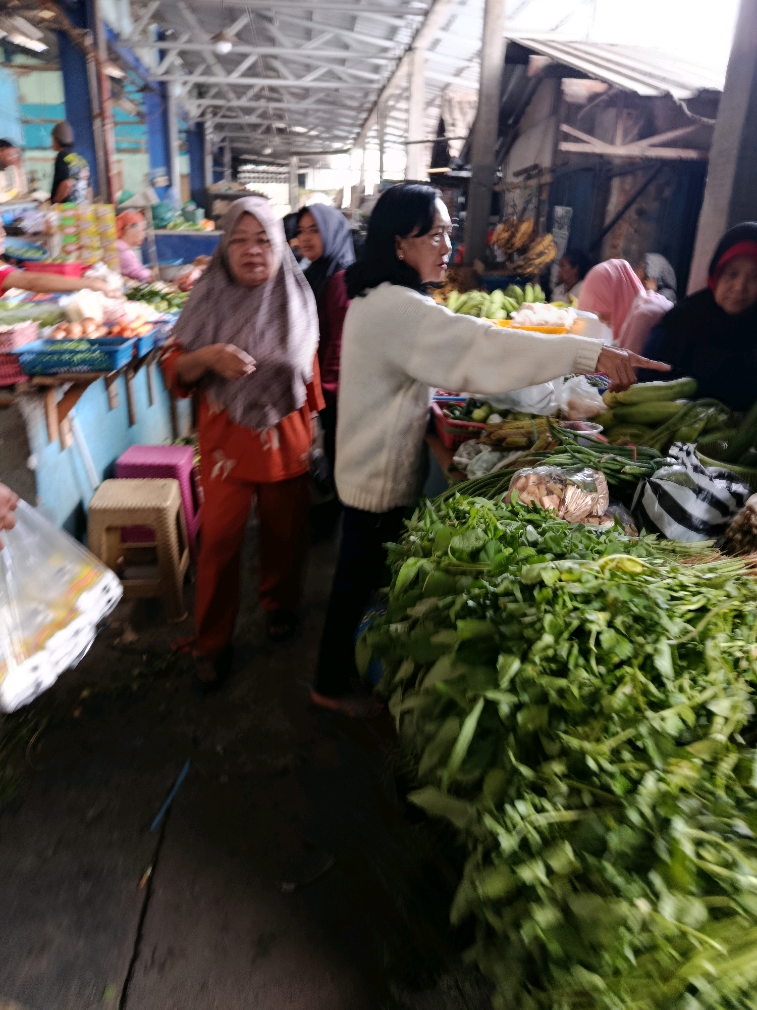 The image depicts a bustling market scene in Bululawang, Indonesia. In the foreground, a vendor with a light-colored sweater points towards a large pile of leafy green vegetables, likely water spinach or a similar local variety, suggesting a transaction or offer. Behind the vendor, a woman in a patterned headscarf and orange attire stands, looking towards the viewer with a neutral expression, possibly a shopper or another vendor. The market appears to be covered with a corrugated metal roof supported by exposed beams, indicating an open-air or semi-open structure typical of Indonesian markets. Stalls with various produce, including bananas and what appear to be root vegetables, are visible in the background, along with other market-goers, some obscured or out of focus. The ground is a rough, paved surface. The overall atmosphere is one of a lively, everyday marketplace.