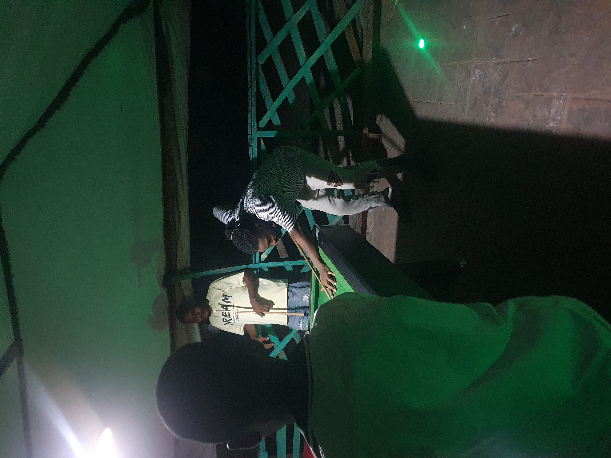 In Lokoja, Nigeria, three individuals are visible in an outdoor, covered area, possibly a patio or veranda, at night. A younger individual, facing away from the camera, wears a green shirt and stands to the left. In the center, a person in a cream-colored t-shirt with "DREAM" printed on it holds a pool cue. To the right, a third person, dressed in a striped shirt and ripped white pants, is actively playing pool, leaning over the green table with cue in hand. The environment features a green-painted lattice fence in the background and a tiled floor. A bright green light emanates from the lower right corner of the frame, casting a strong shadow.