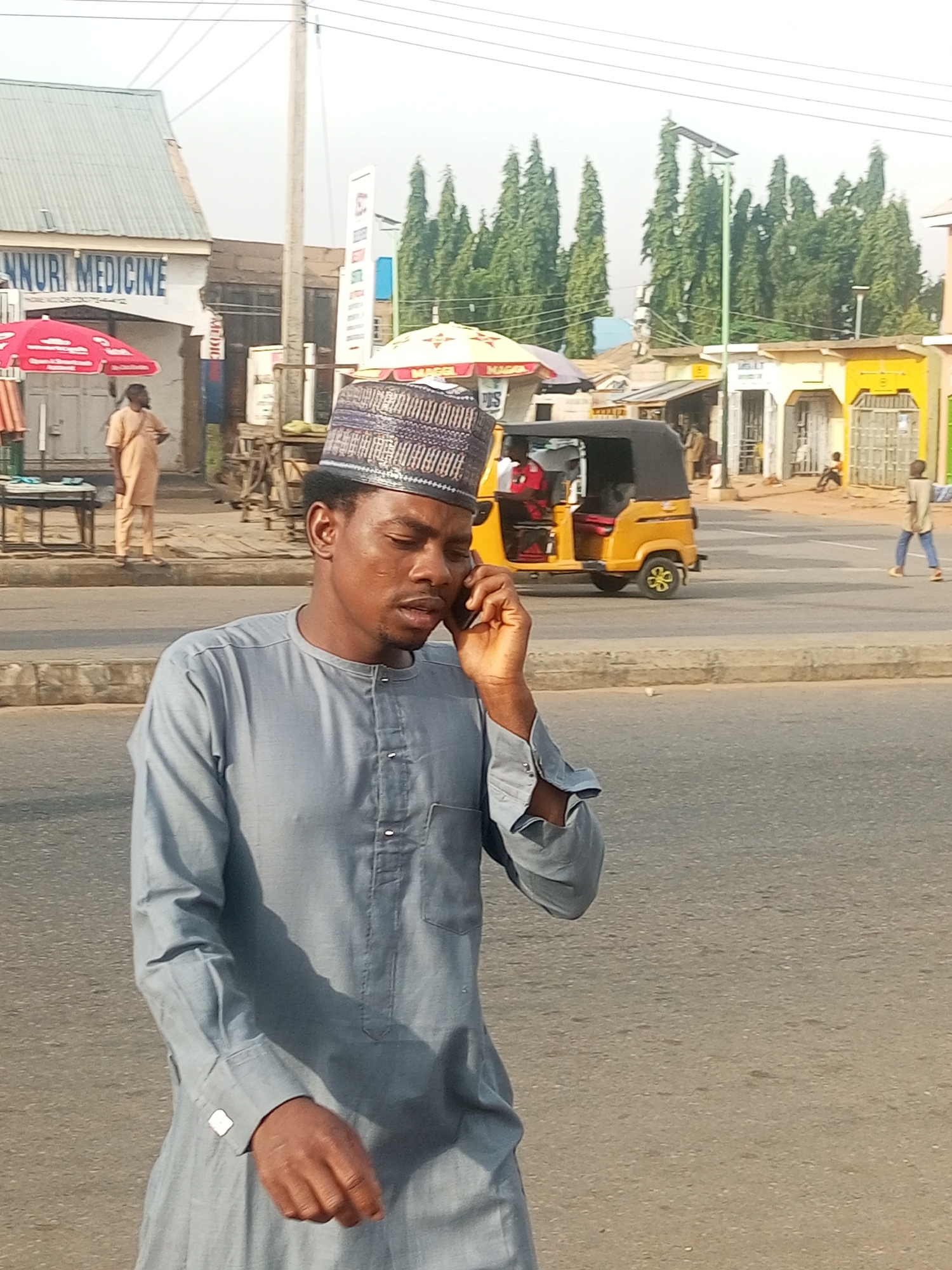 The image captures a street scene with several people and objects. The primary subject is a man in the foreground, wearing a light blue traditional outfit and a patterned cap, speaking on a mobile phone. His expression appears focused. Behind him, a yellow auto-rickshaw with a black canopy is visible in the street.
In the background, there are several buildings with signs, suggesting a commercial area. One sign reads "NNURI MEDICINE". Red and yellow umbrellas are set up outside one of the buildings. Further back, a line of tall, slender trees forms a green backdrop against a hazy sky. The lighting suggests it is daytime, likely late afternoon or early morning due to the soft light. The presence of buildings, street, and people in a busy-looking area indicates an urban or semi-urban setting. Other individuals are present in the background, including a person walking in the distance and another sitting near a building. The overall atmosphere seems like a typical day in a bustling market or street.