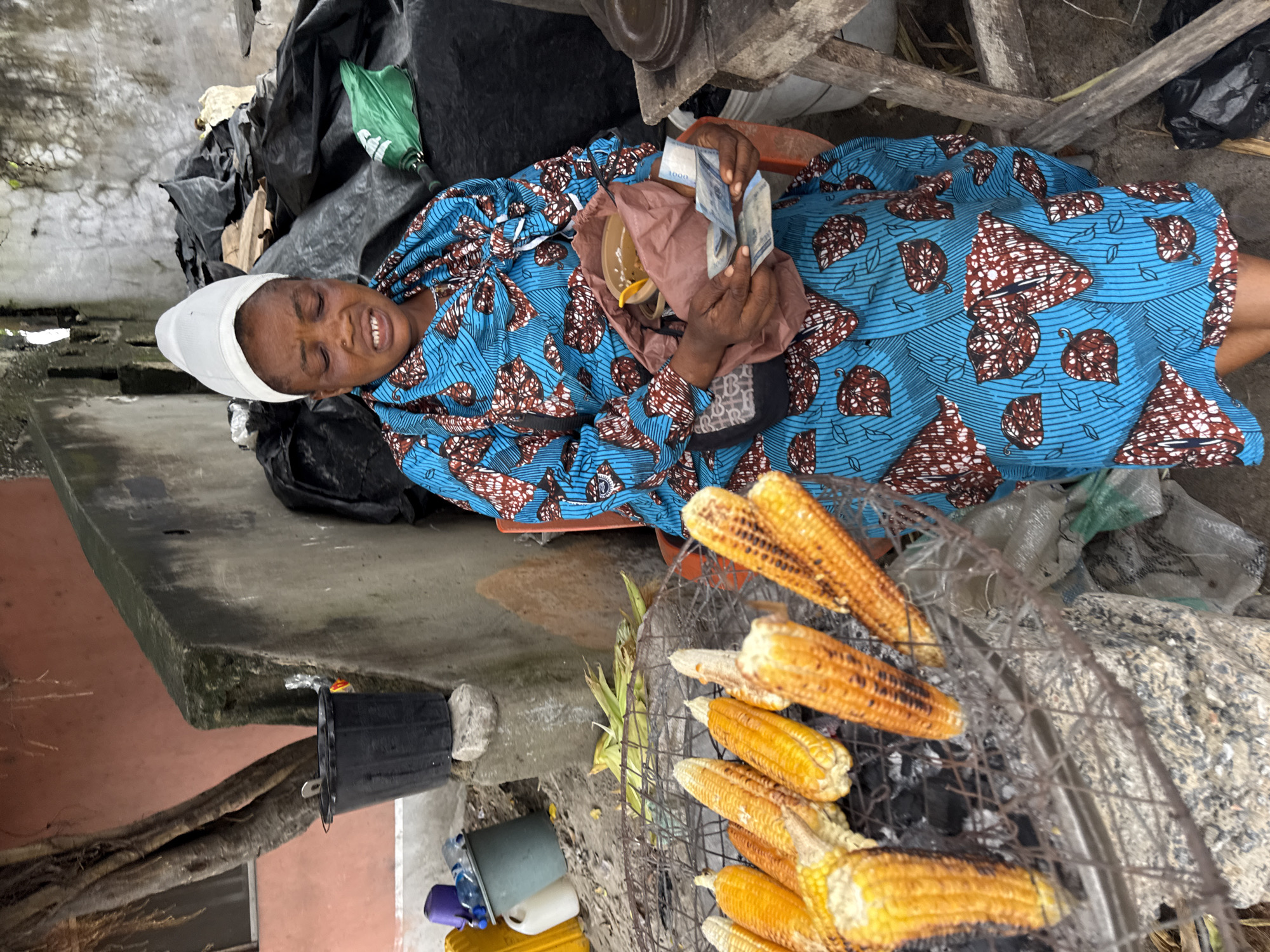 In Ado, Nigeria, a woman wearing a blue and brown patterned dress and a white headscarf sits in front of a grill laden with corn. The corn is golden yellow and appears to be roasting over charcoal. The woman is holding a small pouch and several Nigerian Naira banknotes in her hands, suggesting she is engaging in a transaction. The background shows a rough concrete wall, some black plastic sheeting, and a tree with exposed roots, indicative of an outdoor urban setting. Various objects, including buckets and plastic containers, are scattered around the area. The scene depicts a common street vendor scenario in a Nigerian city.