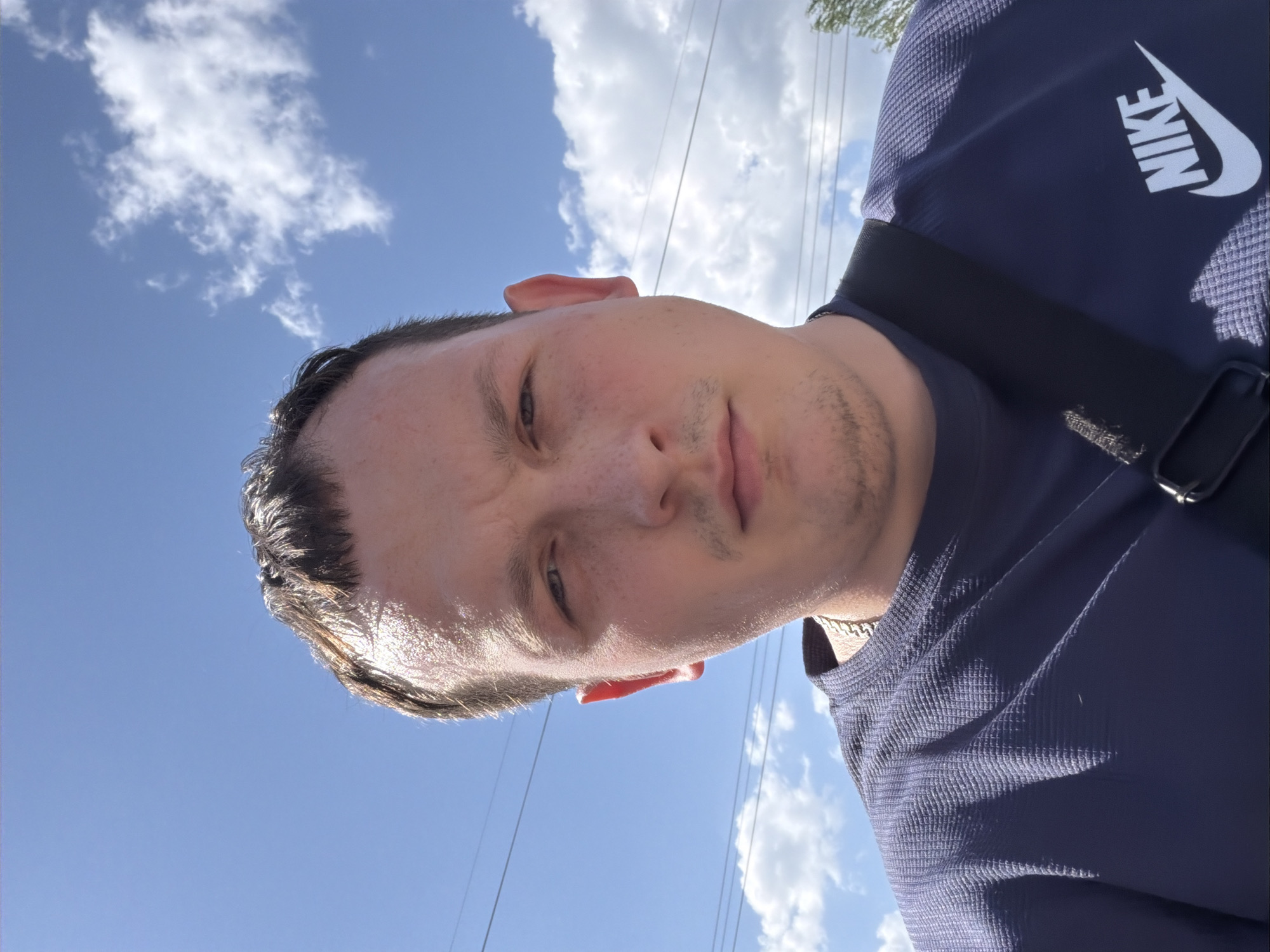 A selfie of a person with short, dark hair and light skin is angled upwards toward a blue sky with scattered clouds. The person is wearing a dark blue Nike shirt and a black strap crosses their shoulder. Overhead power lines are visible in the background. It is impossible to determine the location is Chaikovsky, Russia based on the visual information provided.