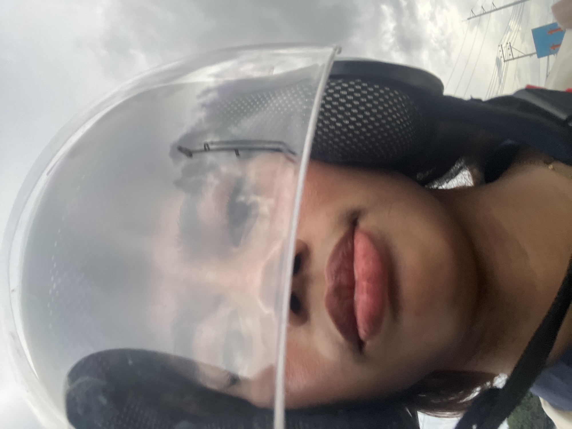 A close-up selfie shows a person wearing a clear helmet. The person's face is visible through the helmet's visor, with their lips and cheeks in focus. The helmet obscures the top of their head and the background. The background appears to be a cloudy sky, possibly with reflections of trees and utility poles visible on the helmet's surface, suggesting an outdoor urban environment consistent with Davao City, Philippines. The person is wearing a blue collared shirt.