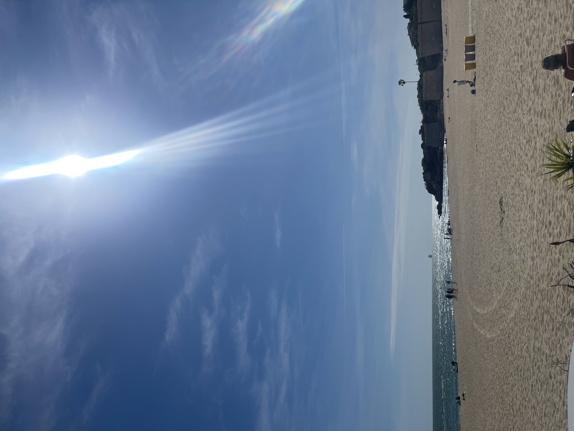 This photo captures a sunlit day at a beach in El Puerto de Santa María, Spain. A wide sandy beach stretches along the right, meeting the ocean on the left. The clear, blue sky is marked by wispy clouds and a bright sun. People are visible along the shore and in the water. Some beach chairs are placed near a series of small structures lining the edge of the beach. The sun's rays are prominent in the sky, creating a vibrant scene.