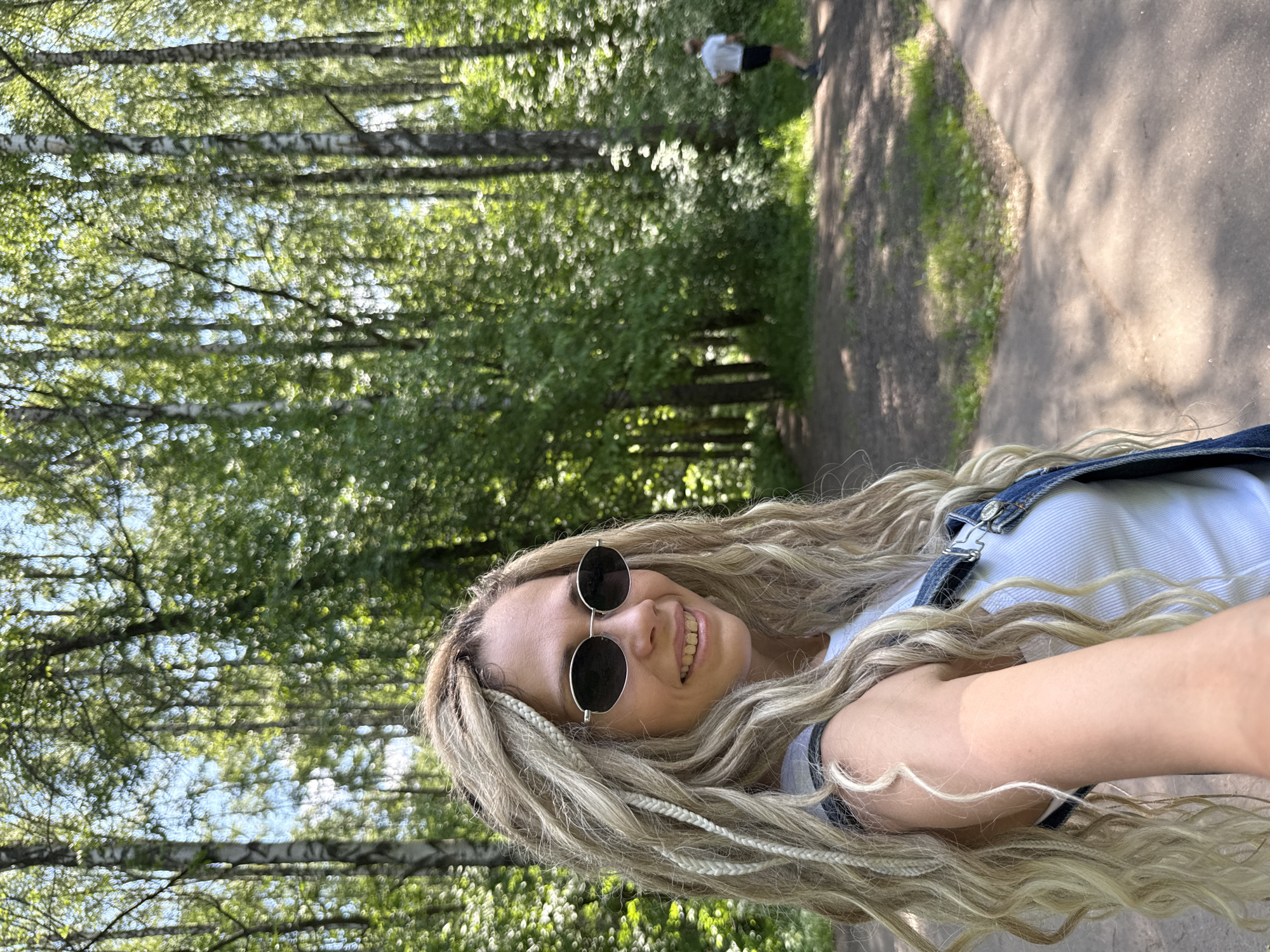 An outdoor, eye-level, medium shot captures a woman with blonde dreadlocks and sunglasses smiling as she takes a selfie in a park. She wears a white tank top and denim overalls. In the background, a man in a white t-shirt and black shorts runs on a paved path. The setting appears to be the city of Rusanovo, Russia, with numerous birch trees and lush green foliage characteristic of a forest park. The lighting suggests it is a sunny day.