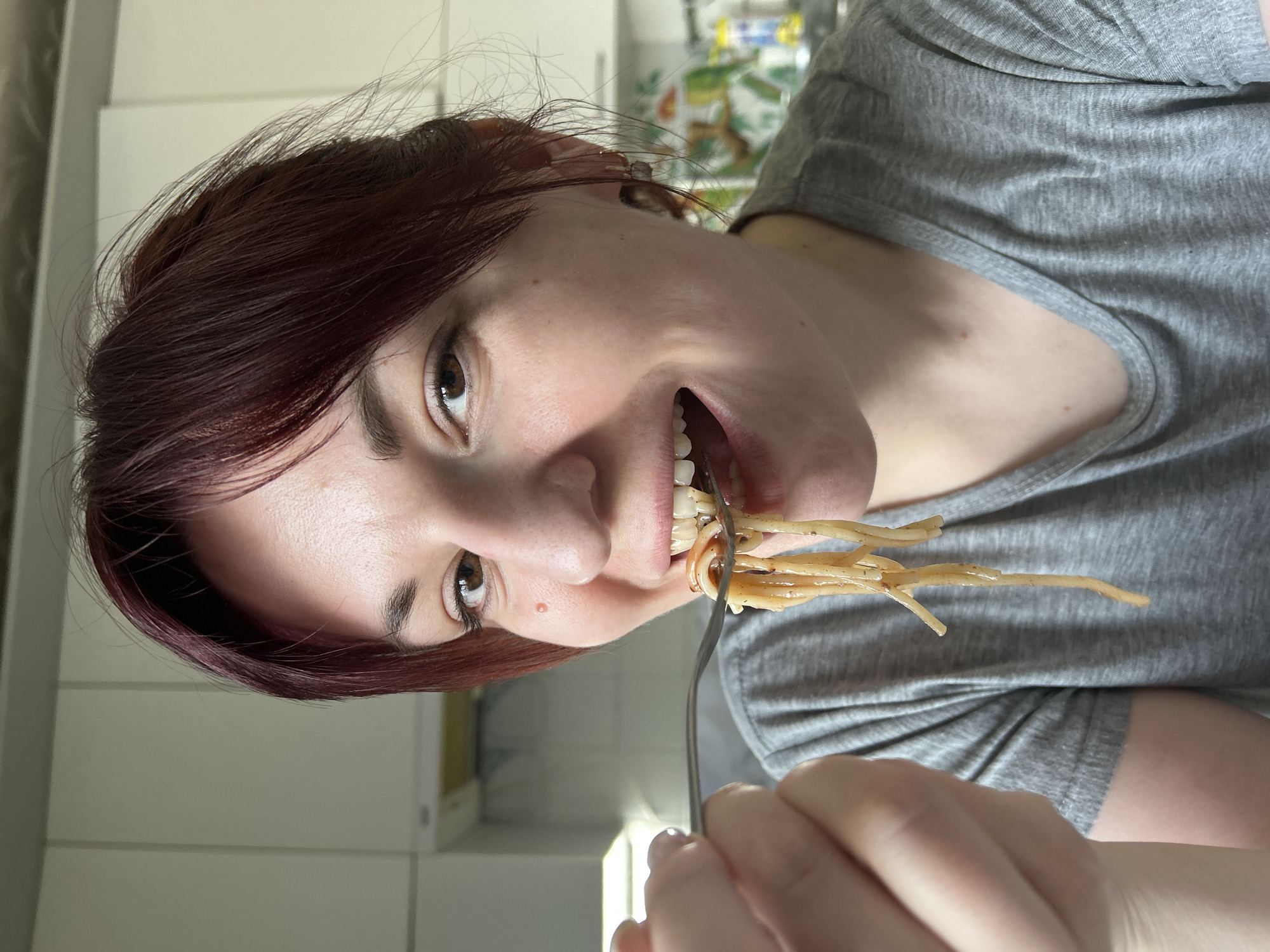 A young woman with reddish-brown hair and light skin is shown eating spaghetti with a fork. She wears a gray t-shirt. The background is blurry but appears to be a kitchen. The location is not discernible as Фоки, Russia, based solely on visual information.