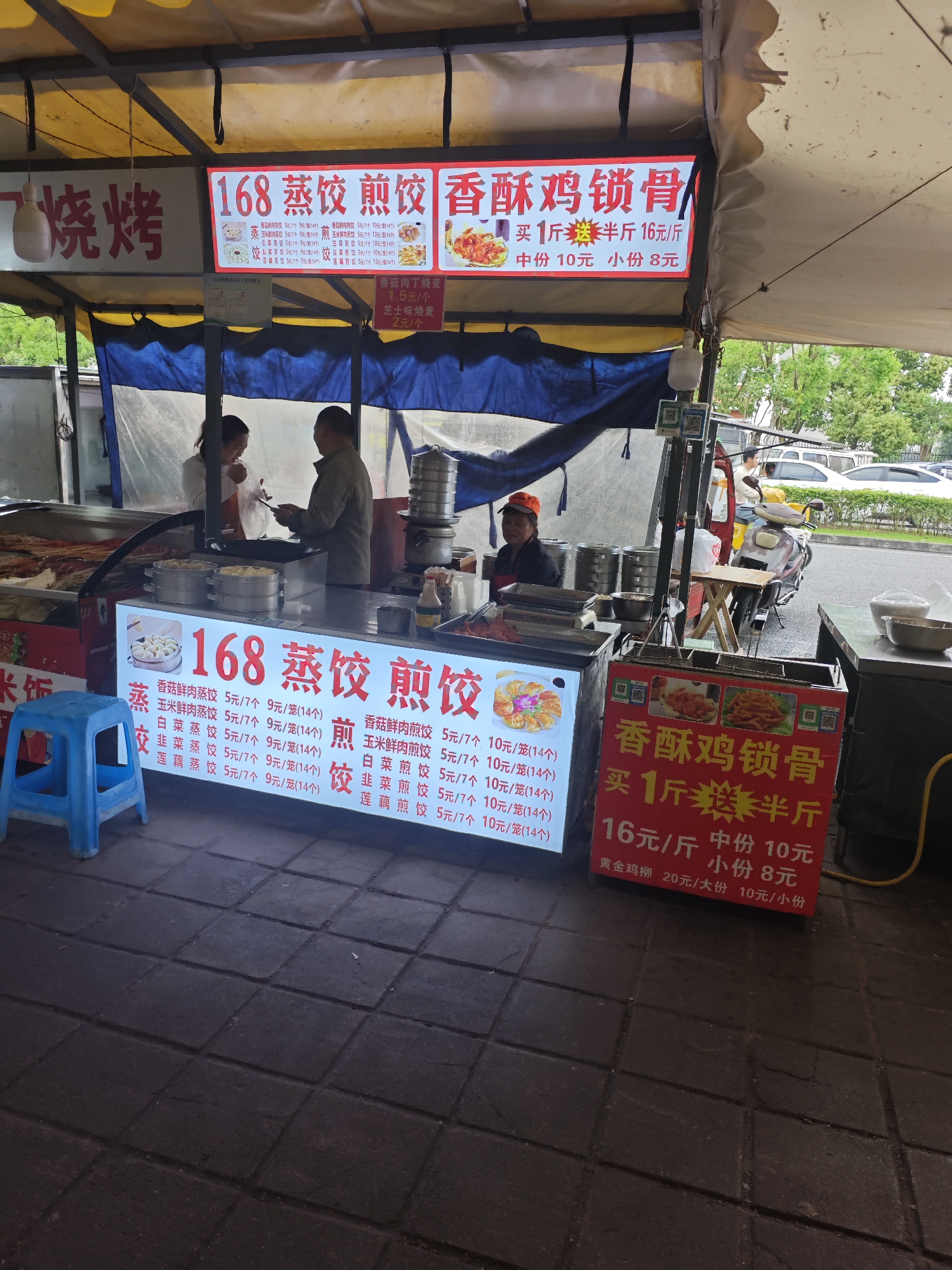 Here's a detailed description of the image:
**Scene:** A street food stall in Shapingba District, China.
**Objects:**
* The stall has a yellow and blue awning, with signs in Chinese advertising "168 Steamed Dumplings, Fried Dumplings, Crispy Chicken Cartilage".
* Signs display prices for various dumpling types (pork, corn, cabbage, leek, lotus root) and the fried chicken.
* Cooking equipment: steamers, fryers, and food containers.
* A small blue plastic stool.
* A motorcycle parked next to the stand.
* Cars parked on the street in the background.
* Serving utensils, small bowls, and condiment bottles.
**People:**
* Three adults, two near the food preparation area and one at the cooking station on the opposite side.
* Two other people are near the motorcycle, appearing to be customers.
**Actions/Interactions:**
* The adults appear to be preparing and serving food to customers.
* Two people stand as customers.
**Environment:**
* The stall is located on a paved area.
* Greenery visible beyond the street suggests a park or residential area.
* The street appears to be a service road, possibly for the building across the parking lot.
**Details:**
* The signs are illuminated, suggesting it's either evening or a dark day.
* QR codes are on the signs, likely for mobile payments.
* Different types of dumplings are on the steamer.
* A variety of fried foods are visible.
**Overall Impression:** The image captures a bustling street food vendor scene in a Chinese city.