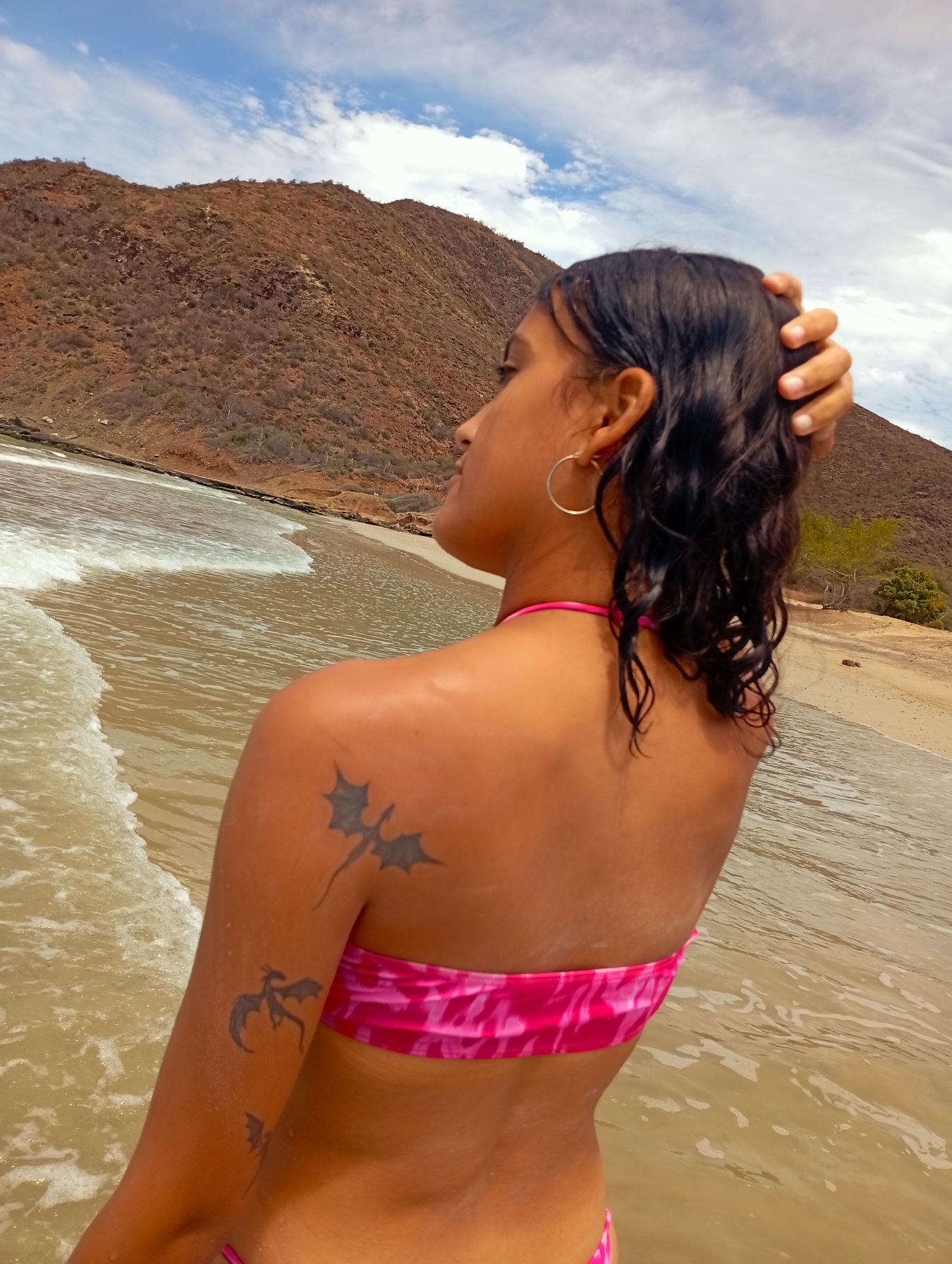 A woman stands in shallow water at a beach in Altagracia, Venezuela. Her back is to the camera, revealing a pink patterned bikini top and tattoos of dragons on her left arm and shoulder. She has dark, wet hair and is wearing hoop earrings. Her right hand is touching her hair. The beach is sandy, and gentle waves are lapping the shore. In the background is a large, dry-looking mountain under a partly cloudy sky.