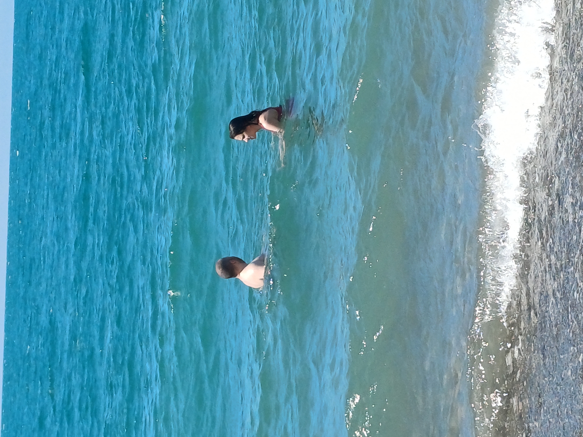 The media file shows two people swimming in the ocean. The water is a vibrant turquoise color, and there are gentle waves. On the right side of the image, a foamy wave is breaking, indicating the edge of the sea. The setting appears to be a beach or a coastline.
One subject is a woman swimming in the water, with her head and shoulders visible. She has dark hair and is wearing a reddish-colored swimsuit. The other subject is a man swimming further away from the camera. Only the back of his head and shoulders are visible, and he appears to be wearing a light-colored garment.
The image is oriented vertically, suggesting it was taken with a phone camera held in portrait mode. The lighting indicates it is daytime, likely sunny, given the bright water and reflections. There are no visible objects other than the people and the water. No text is present in the image. The overall mood seems relaxed and enjoyable, consistent with people swimming in the ocean on a pleasant day. The location context provided is Sochi, Russia.