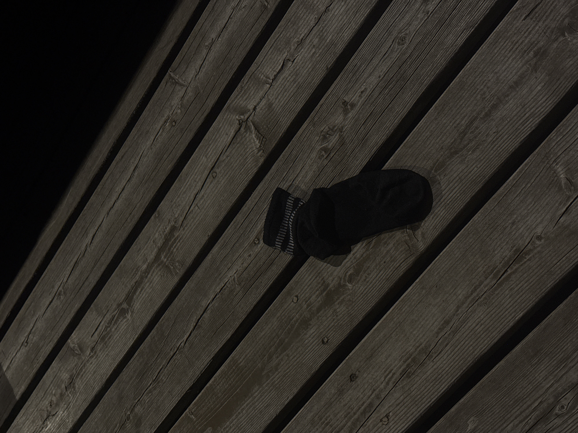 A single black sock with gray stripes lies on a slanted, weathered, dark wooden surface. The surface consists of multiple planks arranged side-by-side. The sock appears crumpled, and the lighting is dim. The scene is likely outdoors at night. The exact location within Nizhny Novgorod, Russia, cannot be determined from the image. No people, actions, or interactions are visible.
