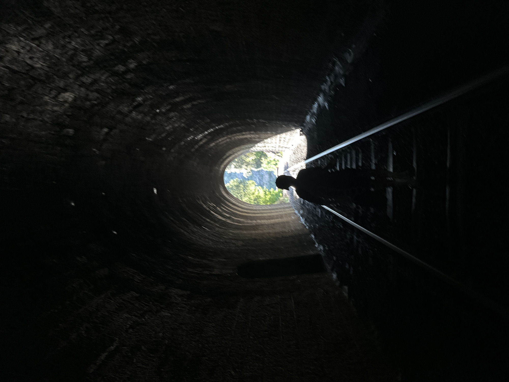 A person walks away from the camera down a railway track inside a dark, arched tunnel. Sunlight streams in from the tunnel's far end, illuminating the silhouette of the person and the tunnel's rough, stone walls. The person appears to be wearing a cap. The tunnel is approximately 150 meters long and is part of the Chemin de Fer du Train Touristique de l'Andouillette, located in Générargues, France.