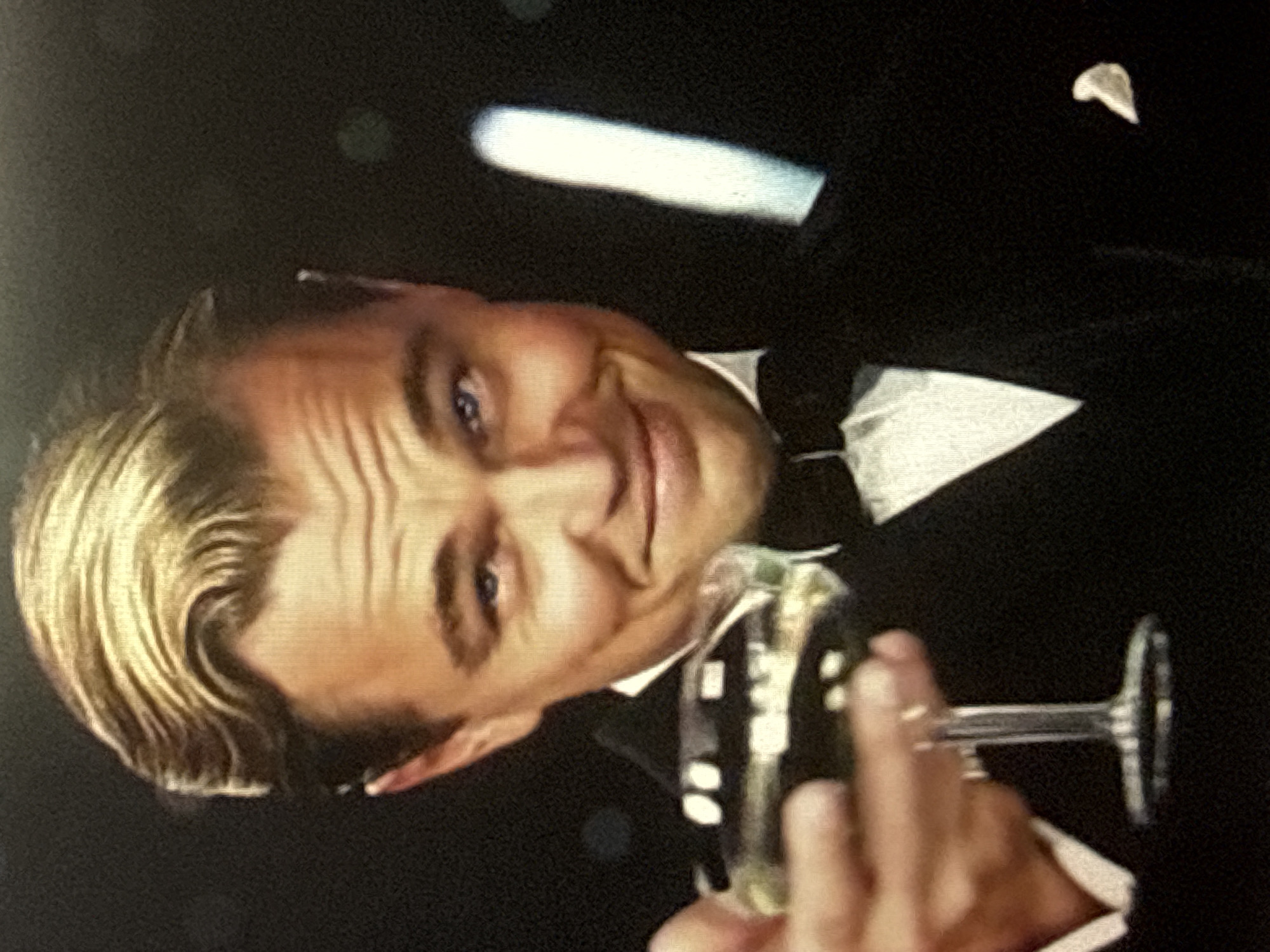 The image is a close-up portrait of Leonardo DiCaprio, styled as Jay Gatsby from the movie "The Great Gatsby". He is wearing a tuxedo with a bow tie and a white shirt. He is holding a champagne flute filled with a light-colored liquid, extending it towards the viewer. His hair is slicked back, and he has a subtle smile. The background is dark with a bokeh effect, suggesting a dimly lit or nocturnal setting. There is no indication of the city of Tbilisi, Georgia, in the image or its context.