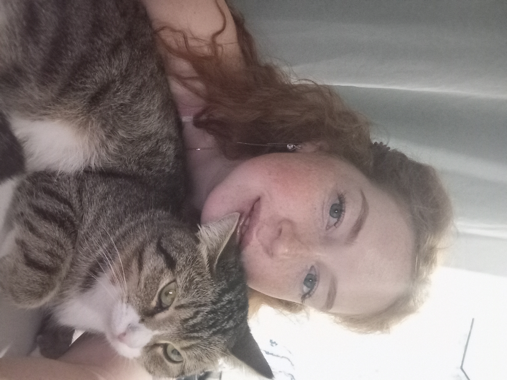 A woman with red hair, light skin, and freckles is lying on her back next to a tabby cat with a white chest and paws. The cat's head is near the woman's face. They are indoors, possibly on a bed, with a white or light-colored sheet partially visible in the background. The exact location within Yurgamysh, Russia, is not determinable from the image.