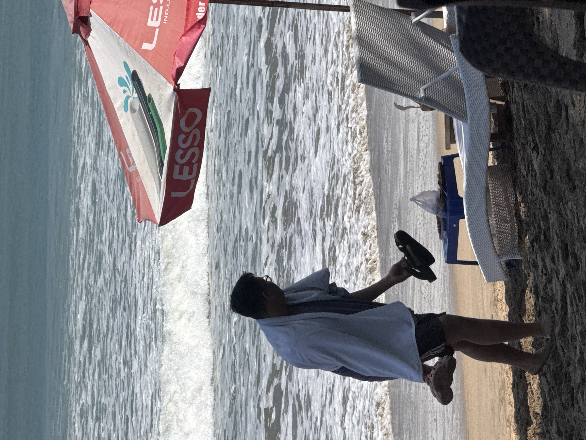 A lone man, draped in a light blue towel, stands on the sandy shore of Kuta Beach, Indonesia. He is holding a pair of flip-flops in each hand, one pair in his right hand and another in his left. The ocean waves are gently rolling onto the beach behind him. A red and white beach umbrella, branded with "LESSO", stands partially visible to his right, providing shade. In the background, a white lounge chair and a small blue table are also present, suggesting a relaxed beachside setting. The scene captures a moment of transition, perhaps the man is about to sit down or leave the beach.