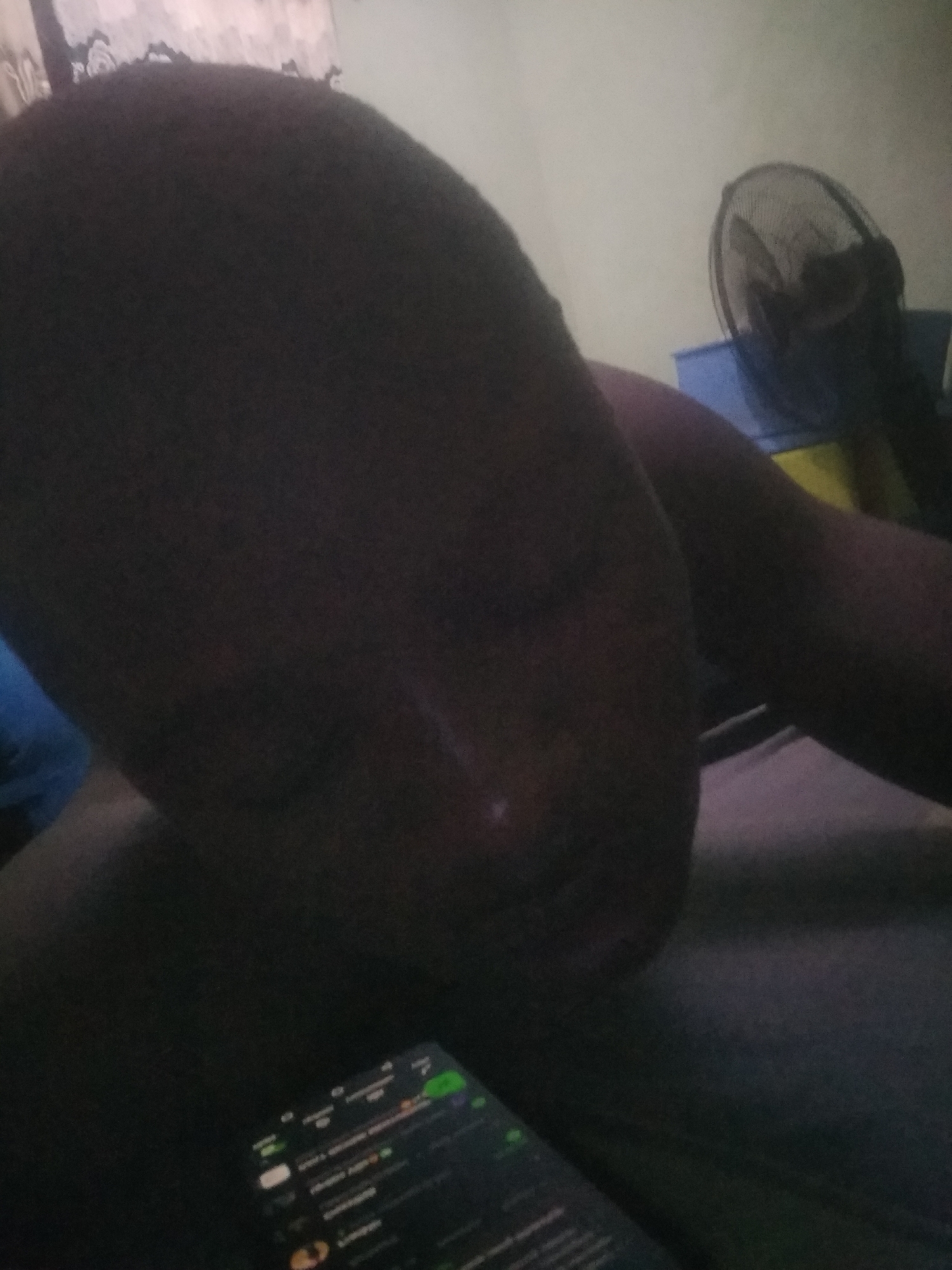 The image shows a person resting or sleeping in a dimly lit indoor environment. The person's face is visible in the foreground, appearing dark due to low light, with eyes seemingly closed. They have dark hair. In front of the person, a smartphone is displayed, showing what appears to be a messaging application with green icons or elements, indicating active chats or notifications. Due to the low light and focus, the specific text on the screen is unreadable, though a few faint characters resembling "Mummy" or "Today" might be perceived. In the background, an electric standing fan is visible against a light-colored wall, with a blue object situated beneath it. The overall atmosphere is quiet and relaxed, suggesting a time of day such as night or early morning, or a room with curtains drawn.