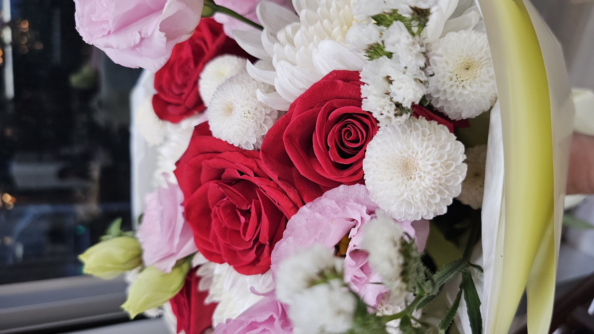 A close-up, vertically oriented, eye-level shot captures a bouquet of vibrant flowers. The bouquet features prominent red roses with deep crimson petals, interspersed with delicate pink roses and clusters of small white flowers, possibly chrysanthemums or asters. Several light green buds, possibly lisianthus or similar, are visible towards the top left. The flowers are arranged in a white container, partially obscured by a light yellow wrapping at the bottom. The background is blurred and dark, suggesting an indoor setting with distant lights, possibly from a cityscape at night. The lighting is soft, highlighting the textures and colors of the flowers. The scene is static, with no discernible action or interaction. There is no visible indication of the location in Asan-si, South Korea.