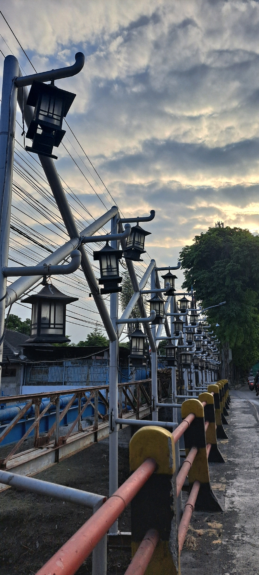 The image shows a row of traditional lantern-style streetlights mounted on metal poles along a pathway next to a bridge. The pathway is bordered by a fence with yellow and black posts and orange and gray railings. A bridge with blue pipes and a rusted metal railing is visible to the left. In the background, there are buildings, trees, and a street with a person on a motorcycle. The sky is cloudy with some sunlight peeking through. This scene is located at the Alun-Alun Pasuruan area in Pasuruan, Indonesia, a popular spot for locals and tourists.