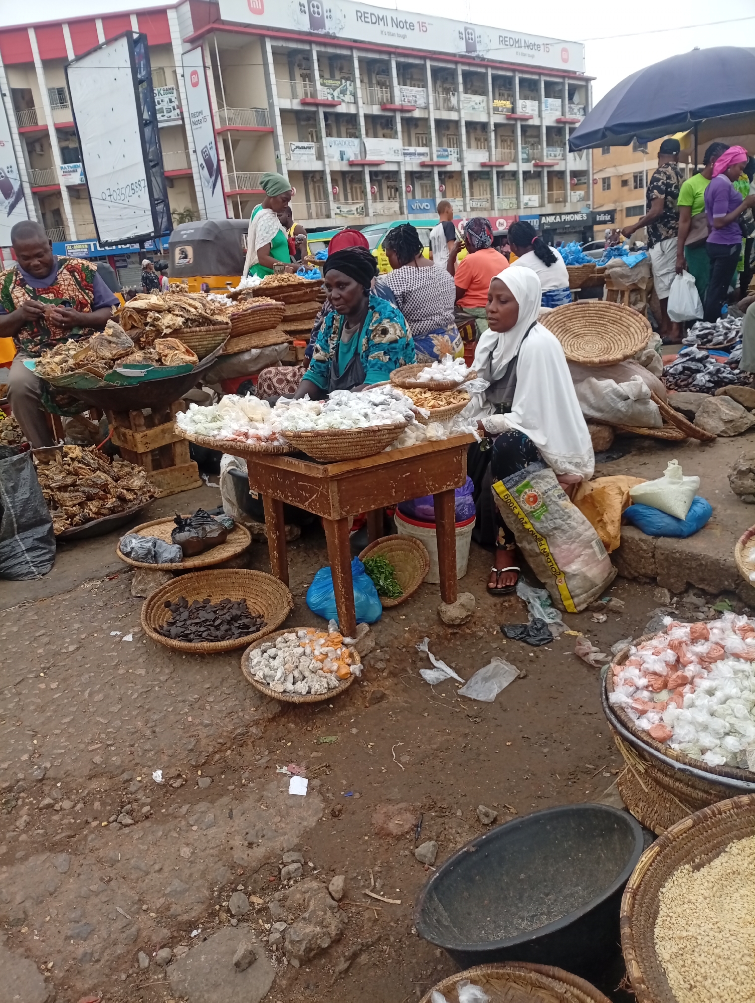 The image portrays a bustling market scene in Maiburiji, Nigeria, marked by vendors and shoppers engaged in transactions. The vendors, predominantly women, are seated at tables or on the ground, displaying their wares in woven baskets. The products include dried goods, possibly fish and spices, packaged in small plastic bags. The ground is unpaved and shows signs of use. In the background, a multi-story building with commercial advertisements for electronic devices and cellular services (Redmi Note 15, Vivo, and Infinix) and business names (AL-AMEEN, ANKA PHONES) is visible. People stand or move about in the background. The presence of a large umbrella suggests an outdoor setting.