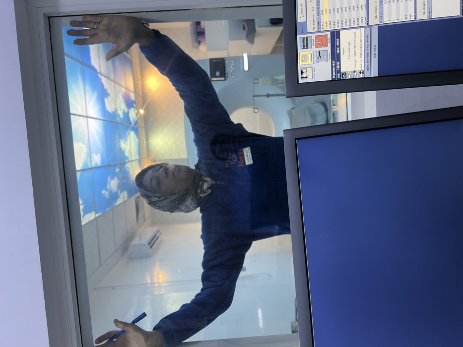 A person wearing a headscarf and blue uniform presses their hands against a glass panel, seemingly from inside a medical or laboratory setting. Reflected in the glass is a room with a simulated sky ceiling, an air conditioner, and medical equipment. Two computer monitors obscure a portion of the scene, one displaying medical data and file names, and the other with a blank blue screen. The person holds a pen in one hand. A name tag on their uniform reads "Nabila".