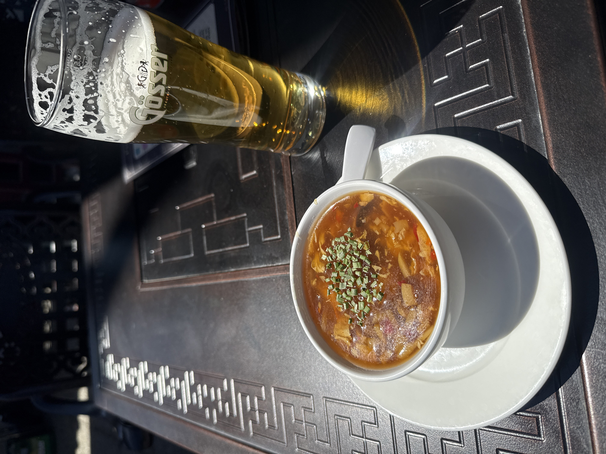 The image presents an overhead view of an outdoor dining setting featuring a glass of beer and a bowl of soup on a dark, patterned table. Bright sunlight casts strong shadows and reflections, indicating a clear and sunny day, likely mid-day or afternoon.
A tall, clear glass of light-colored beer, possibly a lager, occupies the left side of the frame. It has a generous head of white foam clinging to the sides of the glass. The brand name "Gösser" is prominently displayed in white lettering on the glass, along with the word "Brauerei" (Brewery) and the volume marking "0,5 l" (0.5 liters). The sun shining through the beer creates interesting light patterns on the table surface.
To the right, a white ceramic bowl with a handle holds a serving of hot soup. The soup is a rich, reddish-brown color, suggesting an Asian-style soup like hot and sour. It is garnished with finely chopped green herbs, likely chives or scallions, and visible ingredients within the broth include mushrooms and other darker pieces, possibly tofu or meat. The bowl rests on a simple white ceramic saucer.
The table beneath the items is dark, possibly made of metal, and features an intricate embossed geometric pattern that resembles a stylized Greek key or meander design. This pattern adds a decorative element to the surface. Partial views of another table or chair are visible in the blurred background on the left.
Considering the "Gösser" brand beer, which is Austrian, and the provided context, the scene is set in Hainburg an der Donau, Austria. The overall impression is of a casual and enjoyable outdoor meal on a pleasant, sunny day.