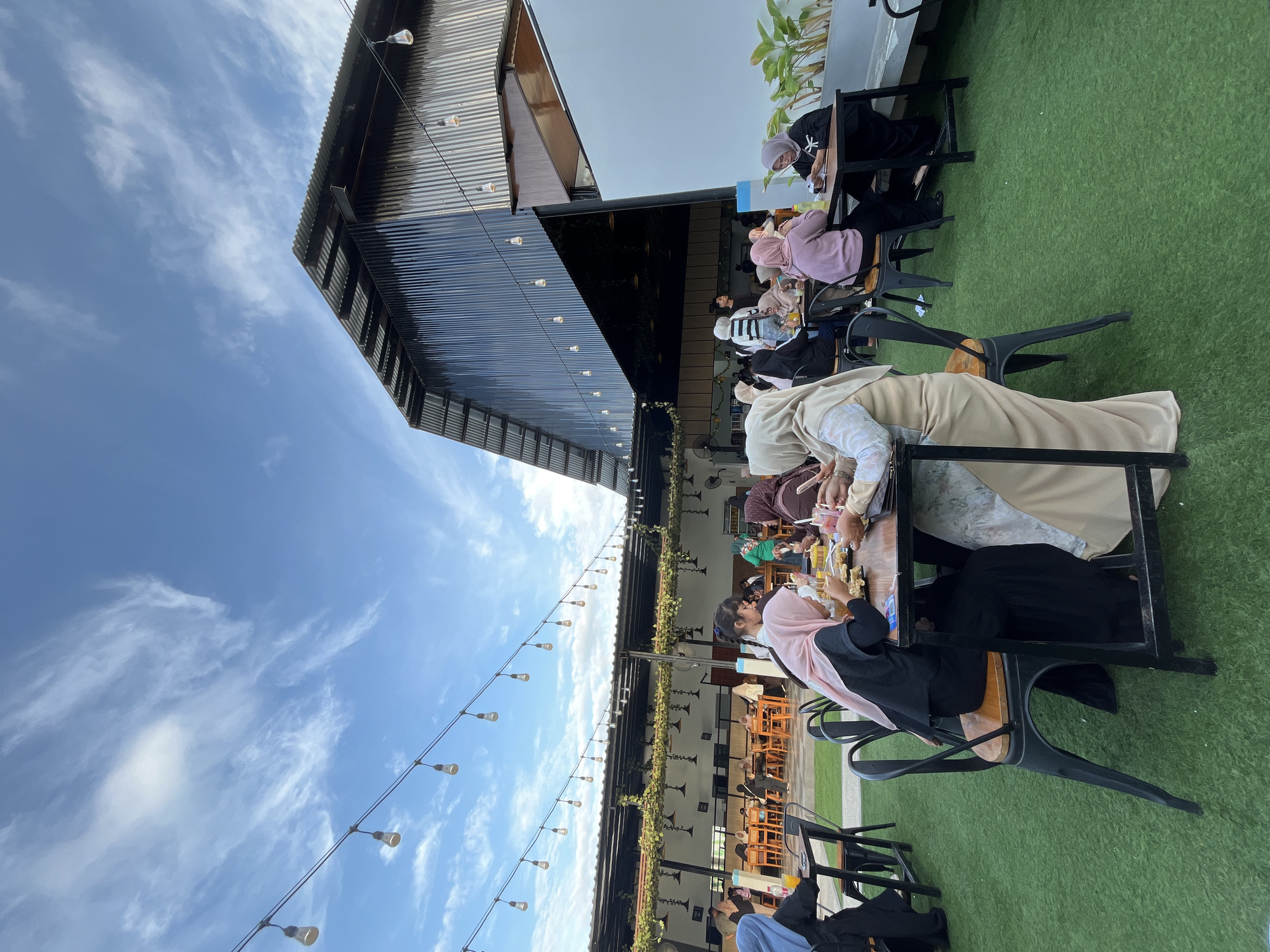 This image captures an outdoor dining area of a restaurant or cafe in Lhokseumawe, Indonesia, under a clear blue sky with scattered white clouds. The ground is covered with green artificial turf.
The scene features a large, dark-colored building with a corrugated metal roof and walls dominating the right side. To the left and extending into the midground, an open-air seating area is visible, covered by a flat roof supported by pillars. This section is decorated with strings of exposed bulb lights and artificial green vines draped along the roof structure.
Approximately 20-30 individuals are present, predominantly women, many of whom are wearing hijabs. In the immediate foreground and midground, multiple groups are seated at black metal tables with wooden tops, using matching chairs. On the right, a group of four women is seated around a table, actively eating and drinking. One woman in a pink hijab and black dress is seen holding a drink and looking down at food on the table. Another woman in a beige hijab is next to her, and across from them, a woman in a black top with a white cross design and a black hijab is looking at something on the table. Various drinks in glasses and plates of food, including small snacks, are on their table. To the left, a woman in a light blue hijab is seated alone at a similar table, drinking from a glass.
In the background, under the covered section, numerous other patrons are seated at wooden tables with orange chairs, engaged in eating, drinking, and social interaction. Some individuals are visible standing or moving within the establishment. On the far right, a white wall section with potted green plants and a low concrete bench is visible. String lights are also strung across the area adjacent to the dark building, with some bulbs appearing illuminated.