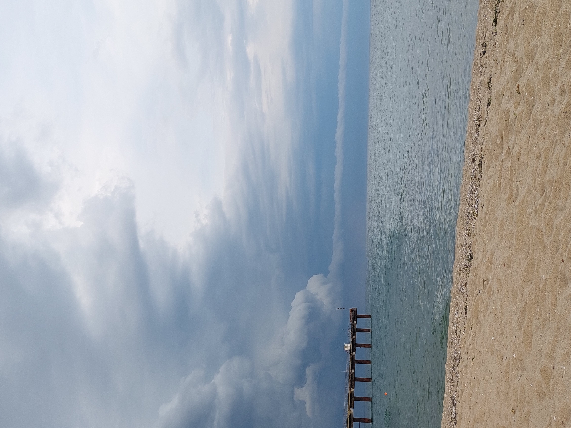 The image depicts a beach scene in Shkorpilovtsi, Bulgaria, characterized by a wide expanse of sand in the foreground, leading to a body of water and a pier extending into it. The sky is filled with dramatic, dark clouds, suggesting an impending storm. The pier, constructed with wooden supports and a weathered deck, has a small, white structure on it. The sea is a calm, muted blue, contrasting with the turbulent sky. The beach itself is composed of fine, light-brown sand, with a scattering of shells and small stones visible along the waterline. There are no people or animals visible in the frame. The overall impression is one of a serene yet imposing natural environment.