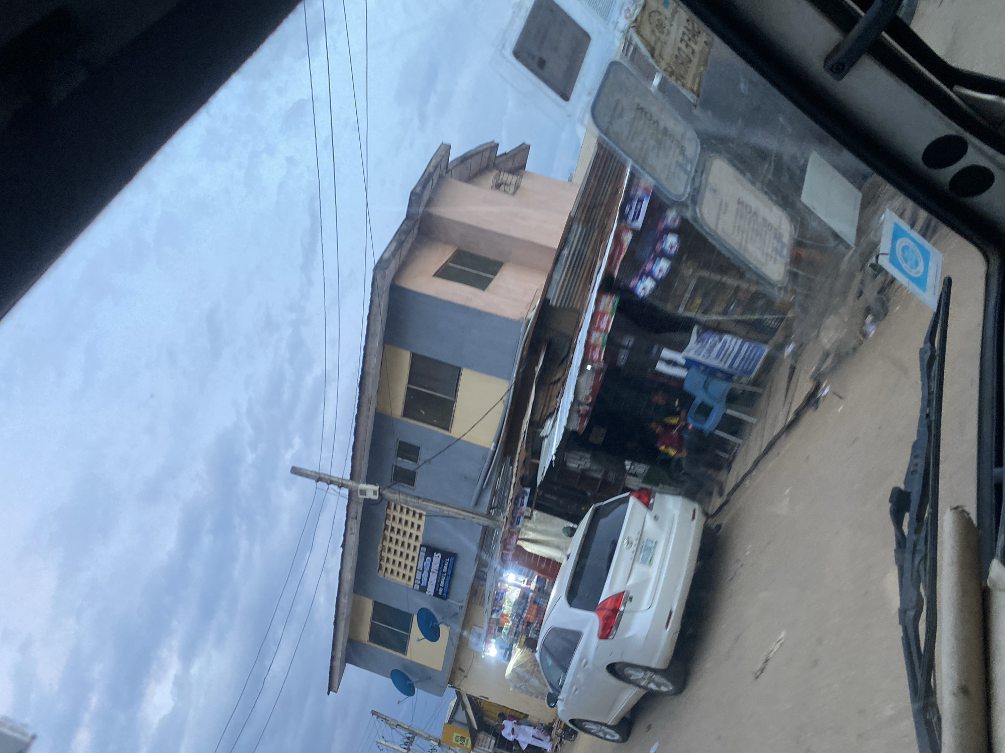 The image depicts a street scene in Ikorodu, Nigeria, captured from inside a vehicle. In the foreground, the dashboard and windshield of the vehicle are visible, with windshield wipers positioned at the bottom. The windshield is adorned with several stickers, some of which are difficult to read.
The main focus of the image is a two-story building on the right side of the street. The upper floor is painted in blue and beige, with windows and two satellite dishes mounted on the exterior. A sign above the entrance reads "SMART PHONES" and "PAY SMALL SMALL." Below the upper floor, the ground floor appears to be a commercial area with shops, characterized by metal awnings and displayed goods, including colorful packaging and bottles.
To the left of the building, a white SUV is parked on the street. Several people are visible in the background, some standing near the building and others seated in blue chairs outside one of the shops. The street appears to be unpaved or dirt road. The sky above is overcast with clouds, suggesting it might be early morning or late afternoon. The overall atmosphere suggests a bustling commercial area in a Nigerian city.