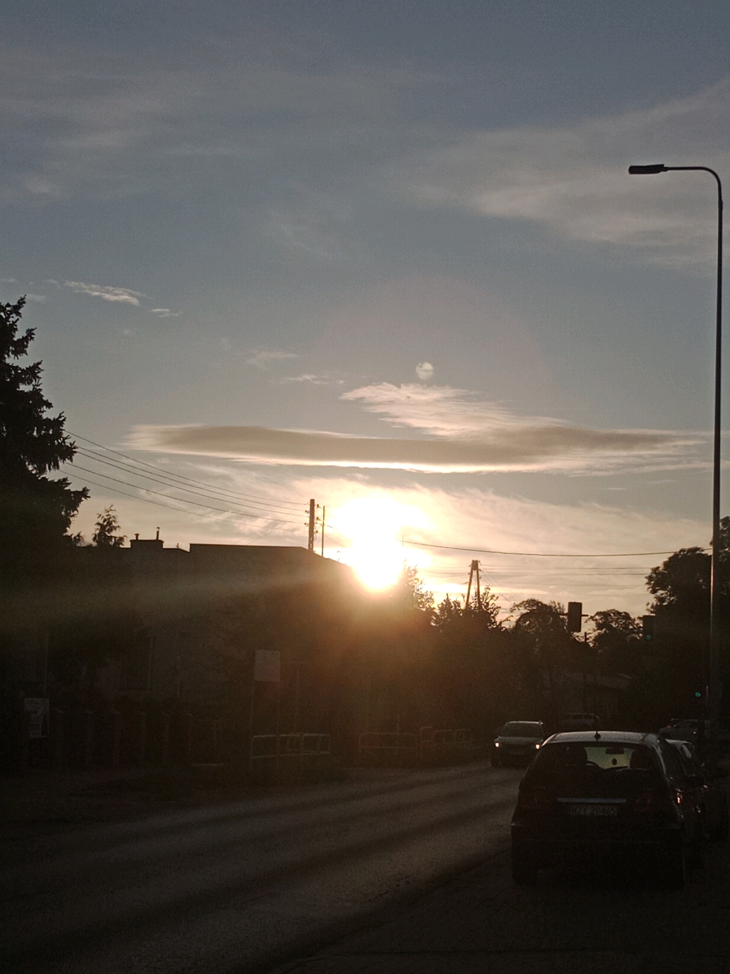 This is a low-angle, eye-level, full shot of a sunset in the city of Bydgoszcz, Poland, specifically along a street lined with buildings and trees. The sun is low on the horizon, casting a warm, orange glow that creates lens flare and illuminates thin, wispy clouds in the sky. A street lamp is visible on the right side of the frame, its silhouette stark against the bright sky. Power lines stretch across the scene, adding to the urban setting. Buildings, some with visible rooftops and windows, are silhouetted against the sunset. Trees, both deciduous and coniferous, frame parts of the image, with one large conifer on the left. The street in the foreground is paved and shows long shadows cast by unseen objects. Several cars are visible on the road, some in motion and some parked. One car in the foreground is dark-colored and appears to be a hatchback, with its license plate partially visible. The overall atmosphere is peaceful and somewhat hazy, typical of a sunset with a touch of atmospheric dust. The presence of streetlights, buildings, and cars clearly indicates an urban environment.
