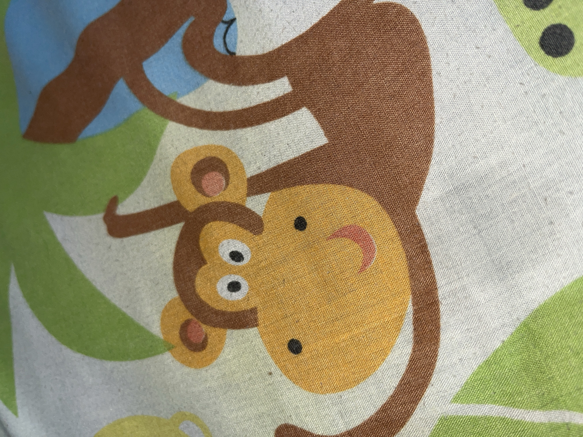 This is a close-up, high-angle, studio shot of a fabric print featuring cartoon monkeys and foliage. The fabric is a light, textured material, likely cotton or a blend. Dominating the foreground is the face of a monkey, rendered in bright yellow and orange, with black eyes and a red smile. Its brown arms and body are visible, with one arm reaching towards the viewer and the other extending upwards. The monkey is depicted swinging from a brown branch, set against a background of stylized green leaves and a blue shape, possibly representing the sky or another element of the jungle scene. The overall impression is of a playful and colorful design. There is no discernible location in Keudematangkuli, Indonesia, as this is an indoor studio shot of fabric.