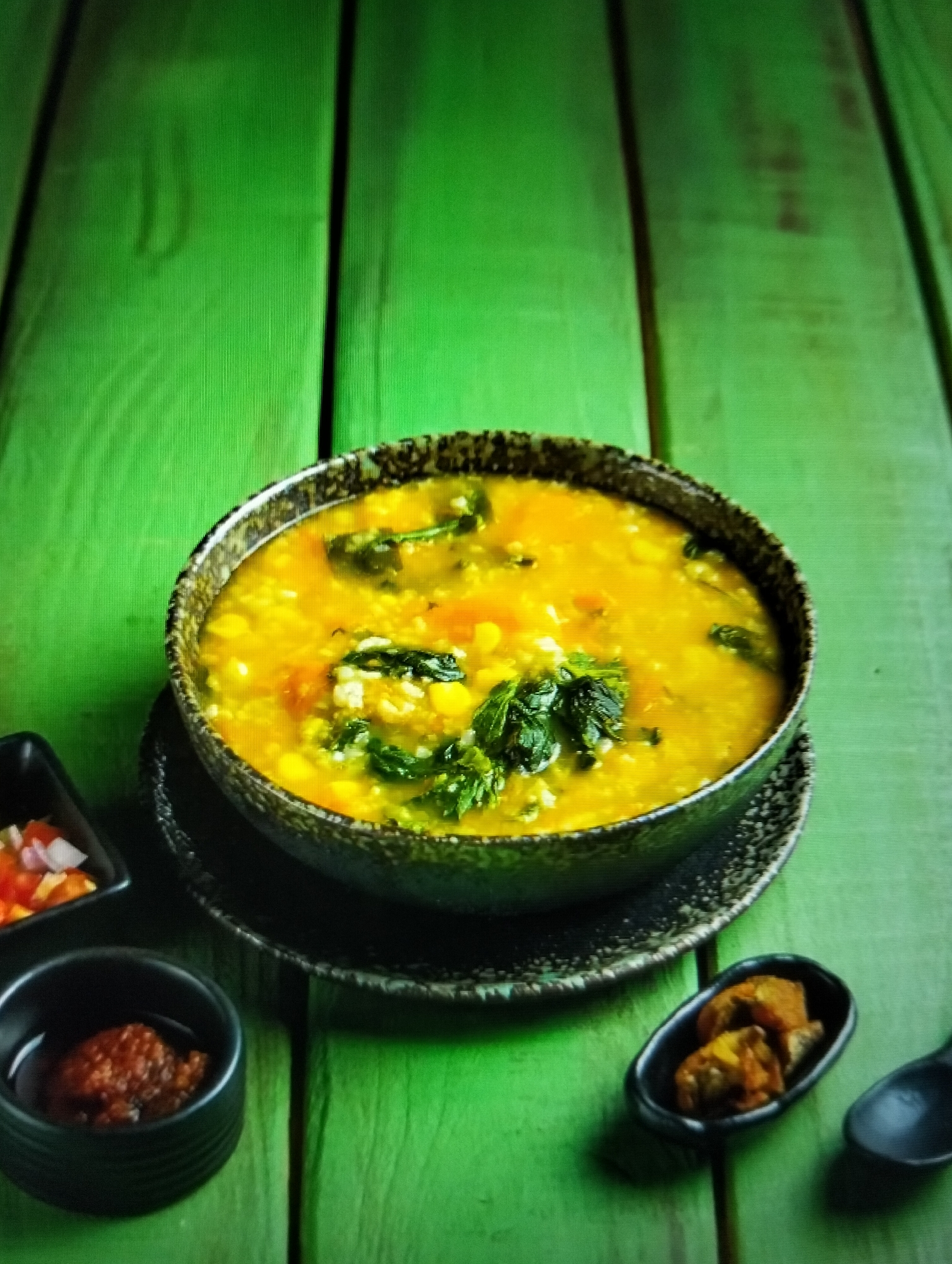 A close-up shot shows a bowl of yellow lentil soup with greens and diced tomatoes, placed on a black saucer. The bowl is centered on a bright green wooden surface. To the left of the bowl, a small black dish contains a red chili paste, and next to it, another small black dish holds chopped tomatoes and onions. To the right of the bowl, a small oval black dish contains what appears to be pieces of fried food, and a black spoon is visible on the far right. The background is out of focus, with the green wooden planks creating a textured backdrop. The lighting is slightly dim, with a noticeable vignette effect. There are no people or animals present in the image. The scene depicts a prepared meal, likely a side dish or accompaniment to the soup.