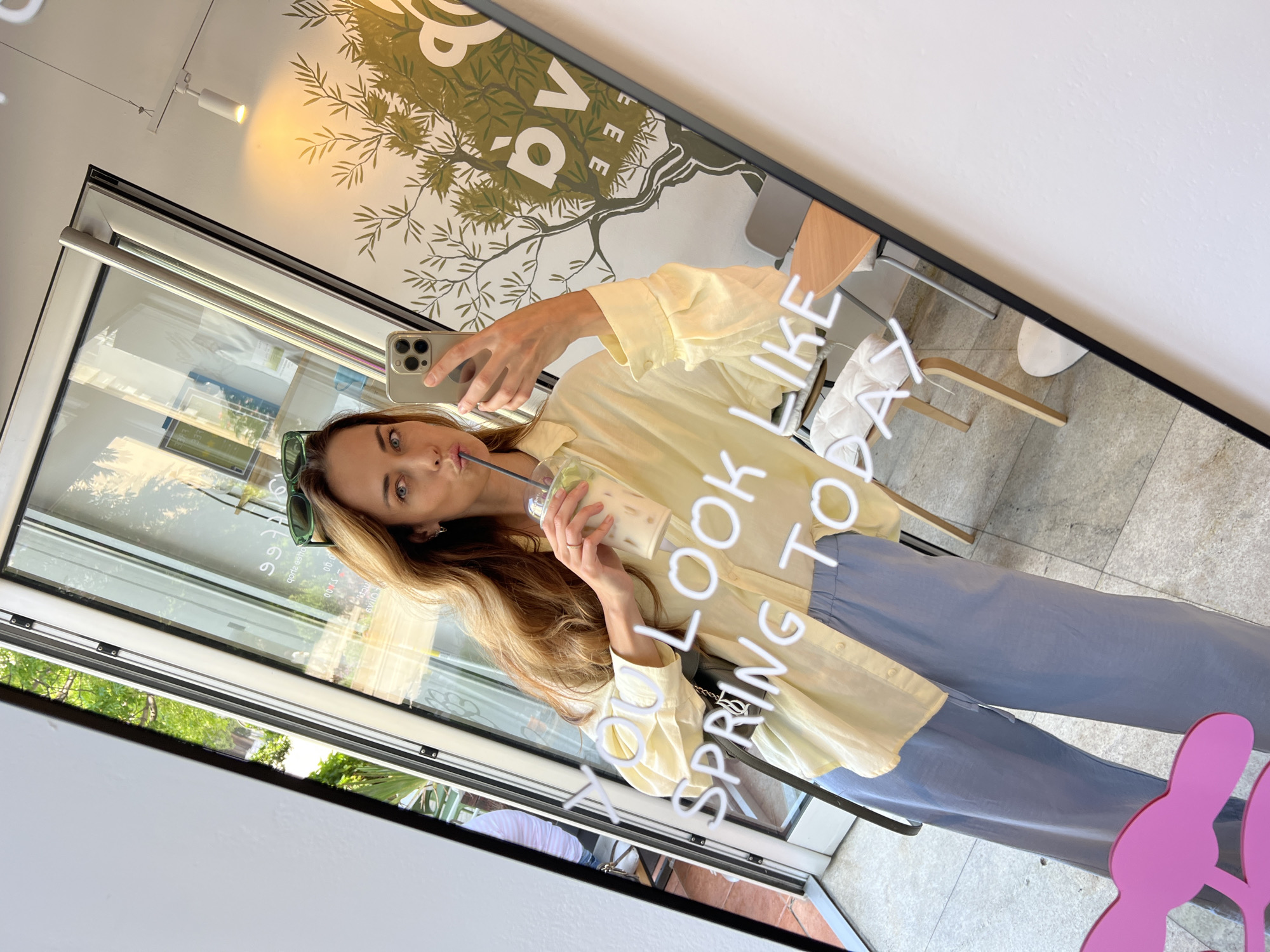 A woman is taking a mirror selfie in a cafe in Bar, Montenegro. She is wearing a yellow oversized shirt and light blue wide-leg pants. She is holding a clear cup with a straw and appears to be drinking from it. Green sunglasses are perched on her head. The mirror reflects the interior of the cafe, which includes chairs, tables, and artwork on the walls. The cafe appears to be modern and airy, with large windows allowing natural light to enter. The text "YOU LOOK LIKE SPRING TODAY" is visible in neon lights on the mirror.