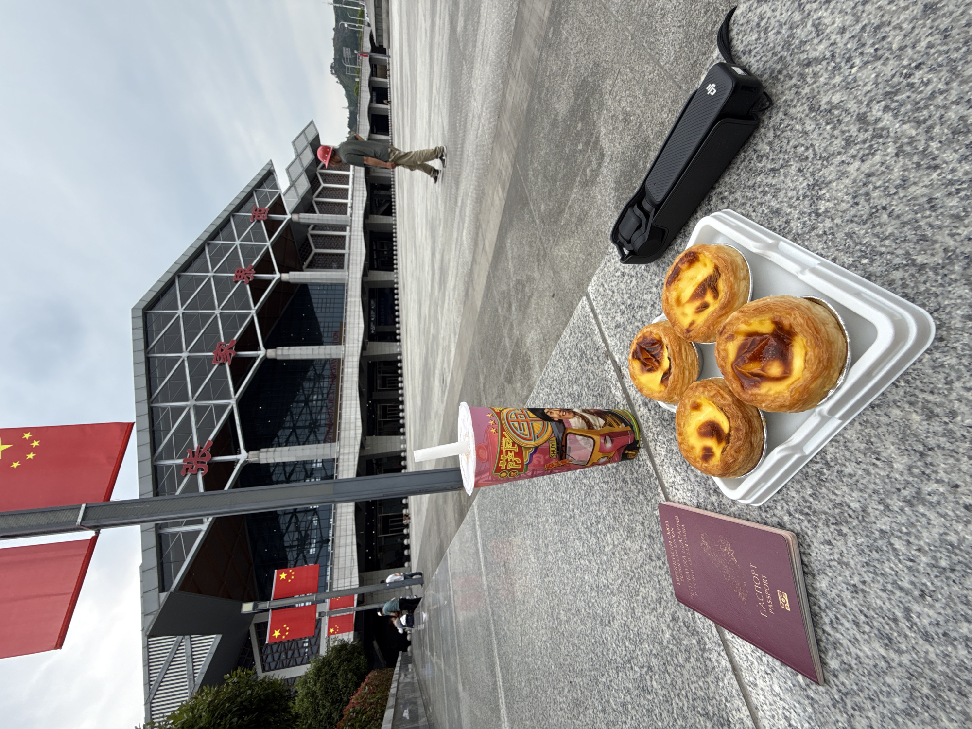 A medium, high-angle shot shows a collection of objects on a grey, tiled surface in Yongding District, China. In the foreground, there is a black DJI Osmo Mobile device, a white styrofoam tray holding four egg tarts, a pink and yellow drink cup with a straw, and a burgundy-colored passport. In the background, a man in a red hat is walking away from the camera near a large building adorned with red Chinese flags. The building features a grid-like design and Chinese characters. The sky is overcast. The environment is clean and urban.