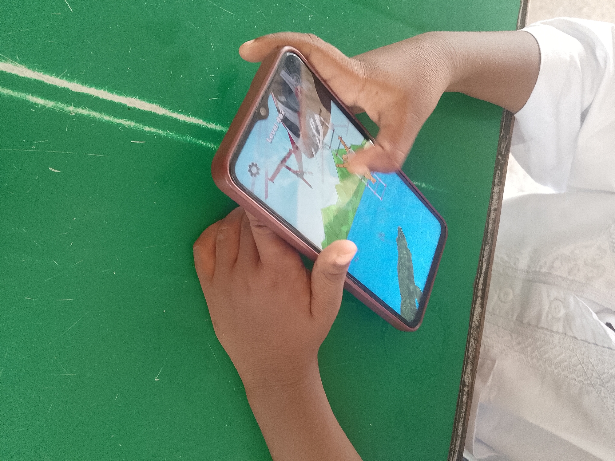 A person is holding a smartphone in their hands, with the screen displaying a game. The phone has a rose-gold case. The person's fingers are touching the screen, indicating interaction with the game. The phone rests on a green, possibly Formica, surface with white lines or scratches. The person is wearing a white garment, and part of it is visible to the right of the phone. Based on the request, this scene is located in Lhokseumawe, Indonesia, although the image does not contain any clear visual indicators of this location.