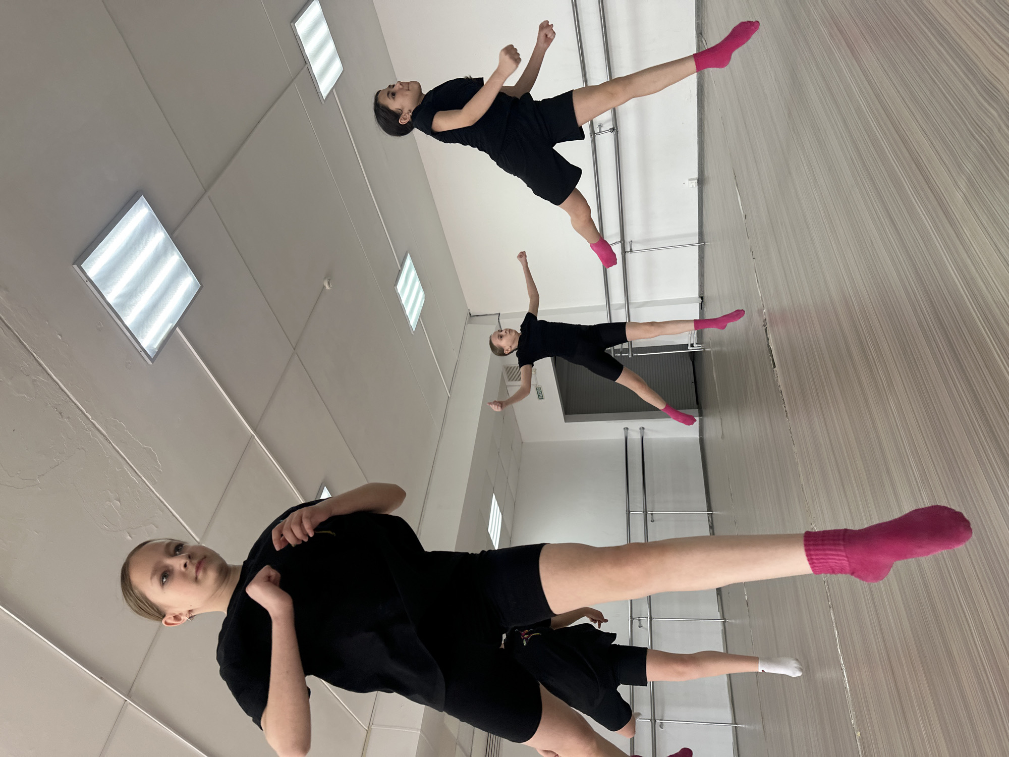 Three young individuals are captured in an indoor dance or gymnastics studio, likely located in Votkinsk, Russia. The image depicts them mid-pose or holding a balance, suggesting a practice session or performance.
The subjects are dressed in uniform black t-shirts and black shorts. The person in the foreground wears bright pink socks, extending her left leg backward while her right arm is raised. A second individual in the middle ground also wears pink socks, striking a similar pose with her right leg extended backward and her left arm raised. The third person, positioned further back, has her left leg extended backward, wearing a white sock, with her arms also raised. Their synchronized poses indicate a focus on form and precision.
The setting is a well-lit studio with light-colored walls and a reflective, possibly wood-patterned, floor. Horizontal ballet barres are visible along one wall in the background, reinforcing the studio environment. The ceiling is a suspended tile ceiling, featuring several rectangular fluorescent light fixtures that provide ample illumination. No specific time of day is discernible due to the indoor artificial lighting, and there is no visible text in the image. The individuals appear focused and engaged in their activity, exhibiting concentration rather than overt emotion.