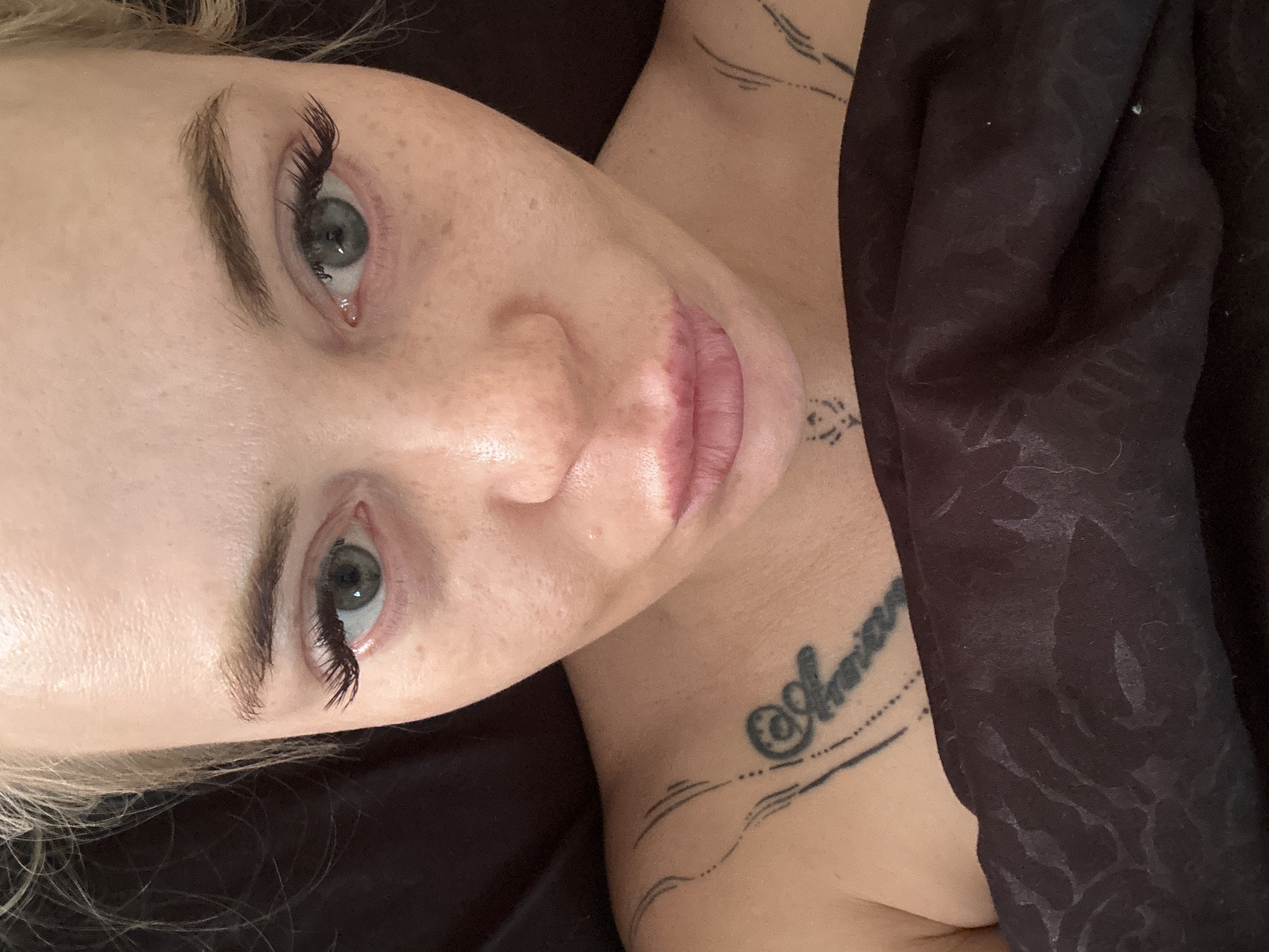 The image is a close-up, overhead shot of a person's face and upper chest, taken indoors. The person has fair skin with freckles, green eyes, and dark blonde hair. They are wearing makeup, including false eyelashes and lip gloss. There are tattoos visible on their chest and neck, including a script tattoo on the chest that appears to read "Annina" and several other linear and dotted designs. The person is covered by a dark, textured blanket that is partially obscuring their lower face and chest. The background is dark and out of focus. The scene is likely a bedroom or other indoor domestic setting, but no specific location within a city can be determined.
