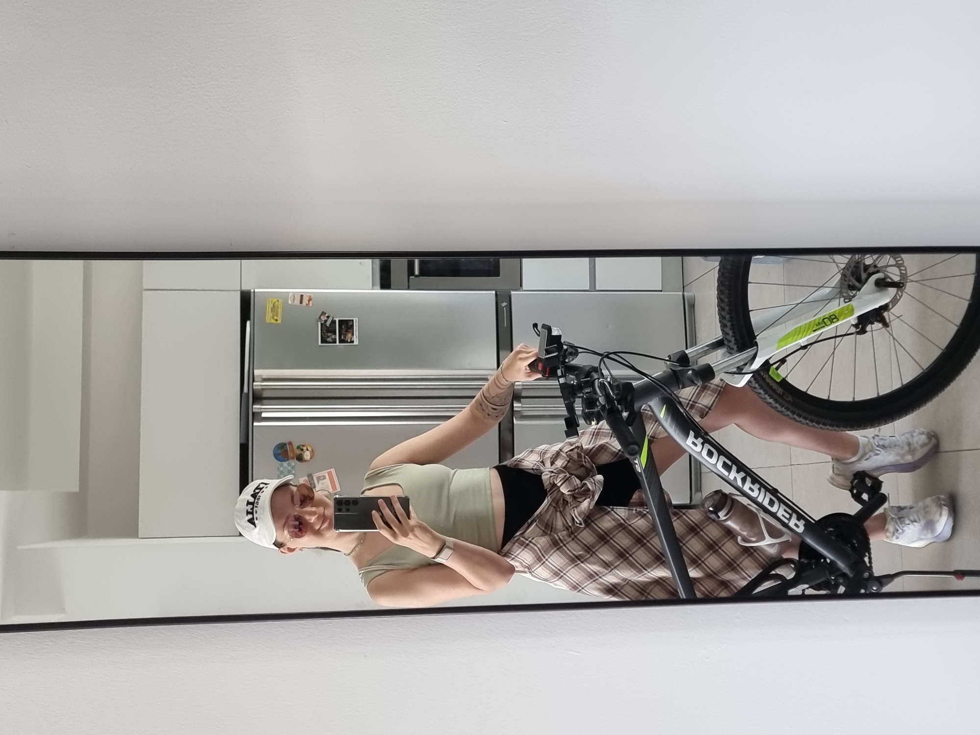 A full-length mirror image captures a woman with a black eye wearing a white baseball cap, a light green tank top, a plaid shirt tied at the waist, black shorts, and white sneakers. She is holding a cell phone, presumably taking the photograph, and posing with a gray and black "ROCKRIDER" mountain bike. In the background, visible through the mirror, is a kitchen area with white cabinets, a stainless steel refrigerator adorned with magnets and photos. Based on prior knowledge of the image source, the location is Ramat Gan, Israel.