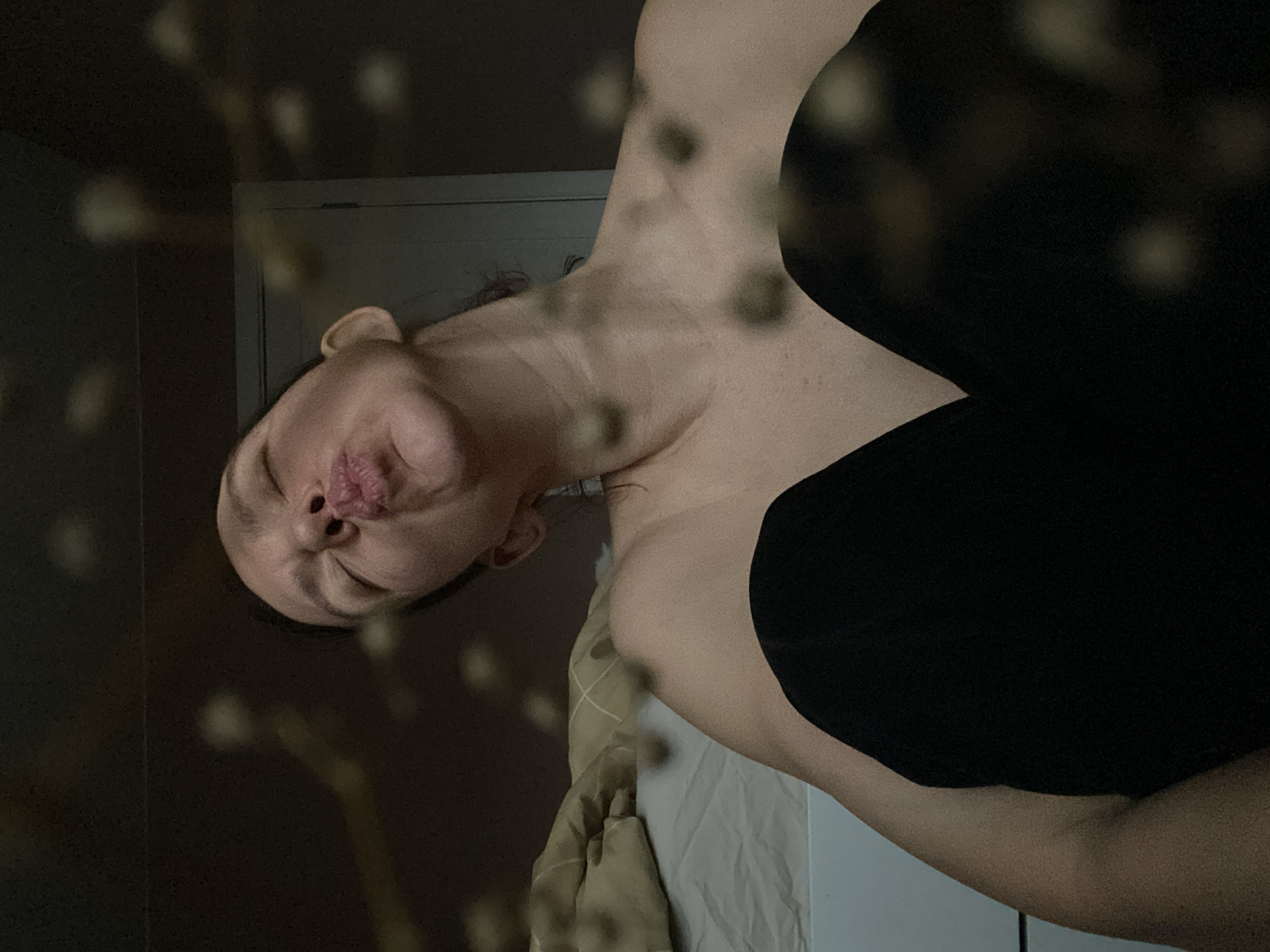 A woman is lying on a bed in what appears to be a bedroom. The room is dimly lit. The woman is wearing black clothing and is turned sideways to the camera. Her face is puckered as if she is posing for a kiss. A beige blanket is visible. The location cannot be determined to be in Chaikovsky, Russia from visual data alone.