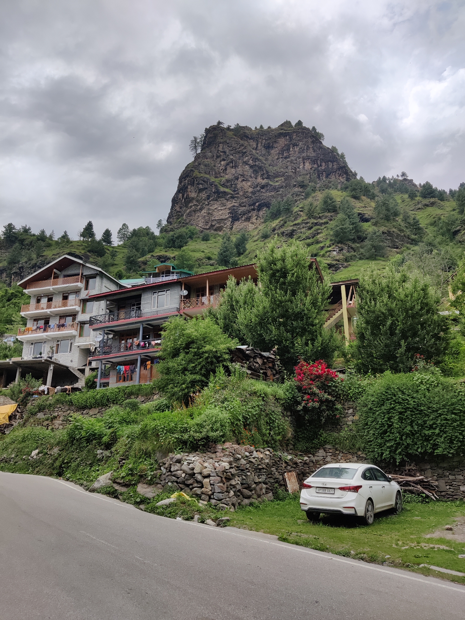 The image depicts a scene in the city of Palchan, India, featuring a road, buildings, and a mountainous landscape. In the foreground, a paved road curves to the left. To the right of the road, a white sedan is parked on a grassy area. Behind the car, a stone wall supports a steep, green hillside.
Further up the hillside are several multi-story buildings constructed with concrete and wood accents. These buildings appear to be houses or guesthouses, with balconies visible and laundry hanging from some of them. Lush green trees and vegetation cover the slopes surrounding the buildings.
Dominating the background is a large, rocky mountain face with sparse trees growing on its slopes and summit. The sky is overcast with heavy, grey clouds. The overall impression is of a rural or semi-urban environment nestled within a mountainous region, likely under a cloudy sky.